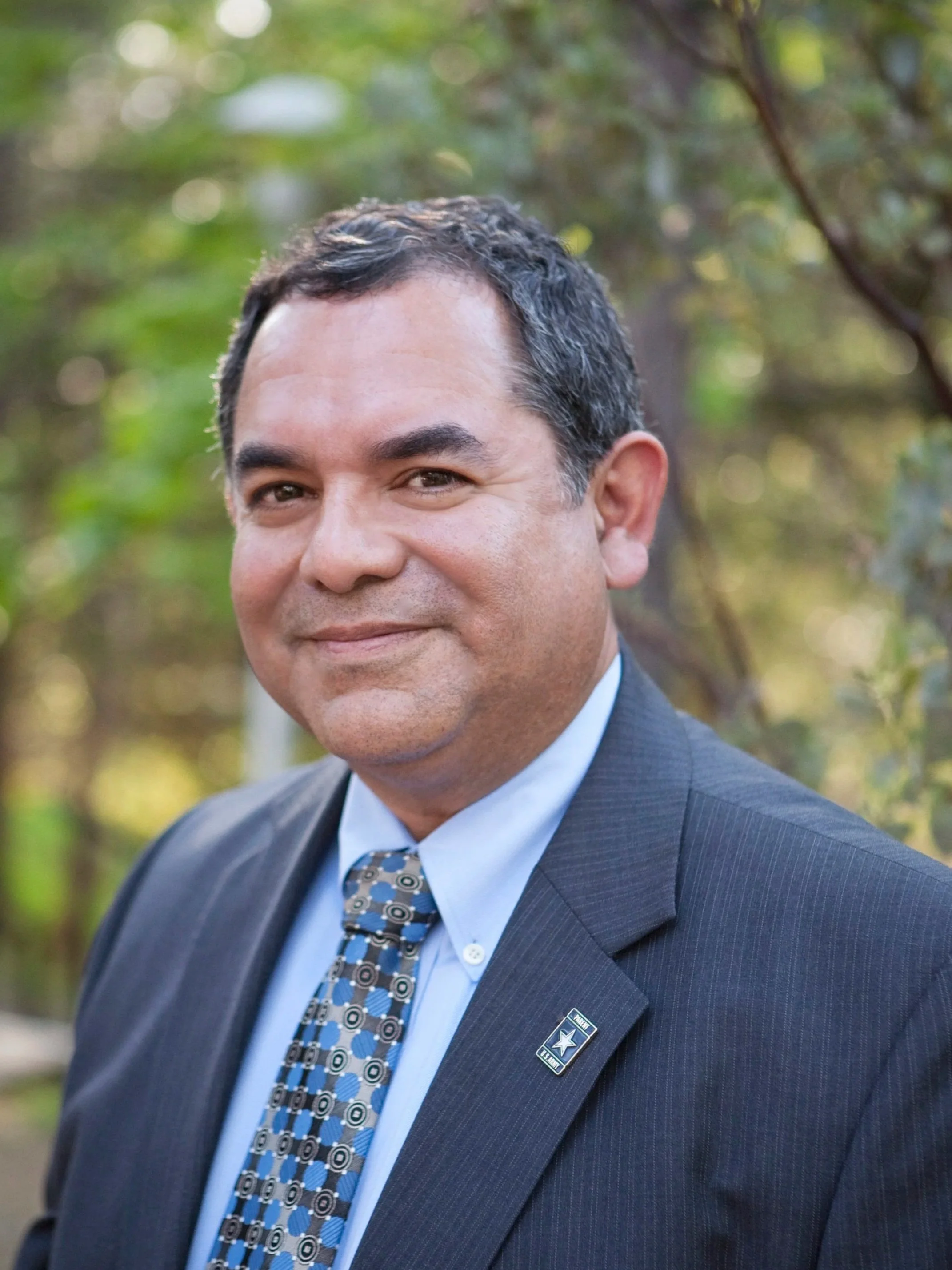 A man with dark hair and tan skin wearing a dark pinstripe suit, blue dress shirt, and patterned tie, standing outdoors with blurry trees in the background.
