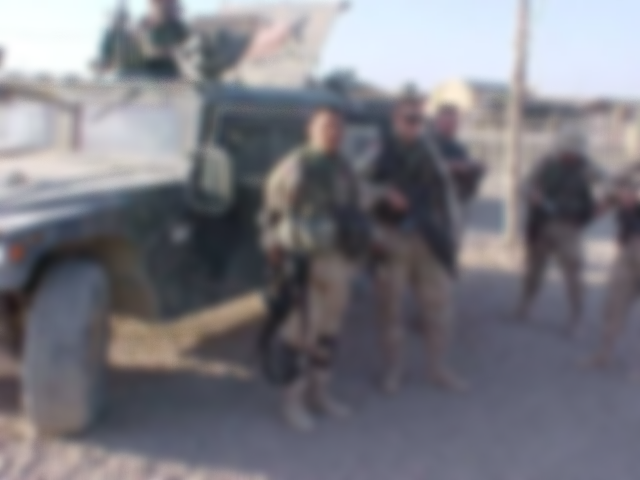 Four soldiers standing in front of a military vehicle in a rural area.