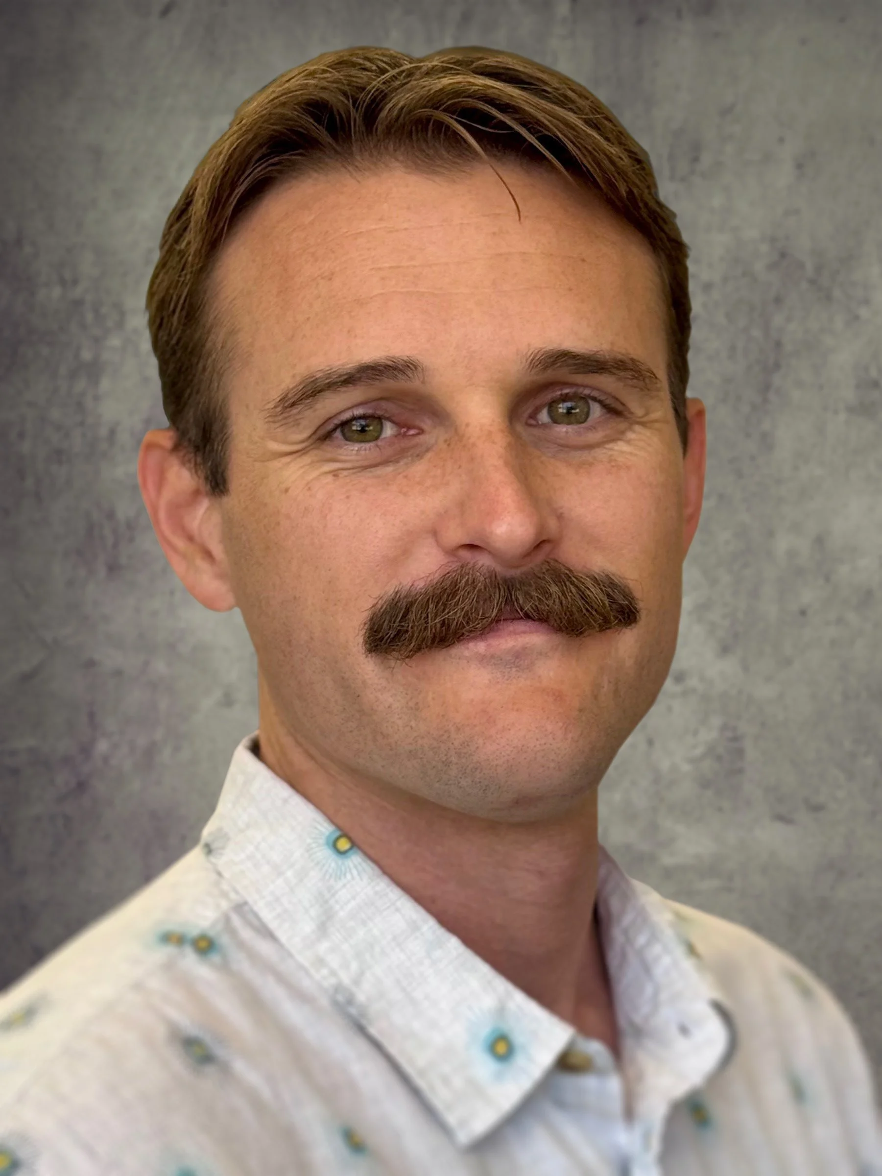 Close-up of Austin Bowman, a man with light skin, brown hair, hazel eyes, and a mustache wearing a light-colored patterned shirt, standing in front of a gray textured background.