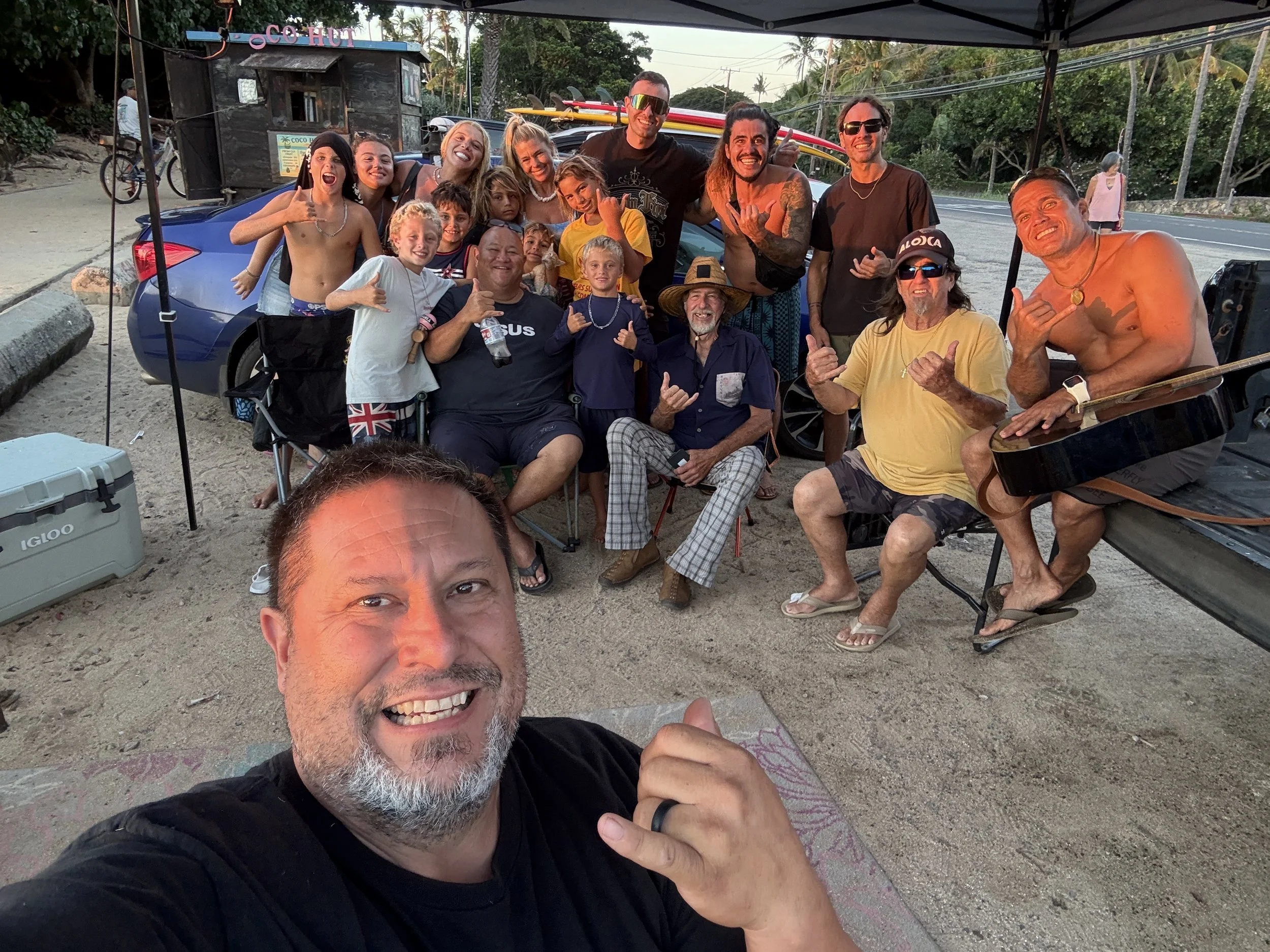 A group of people, including children and adults, smiling and posing for a selfie outdoors near a car with surfboards on top. They are under a canopy with trees and a street in the background.