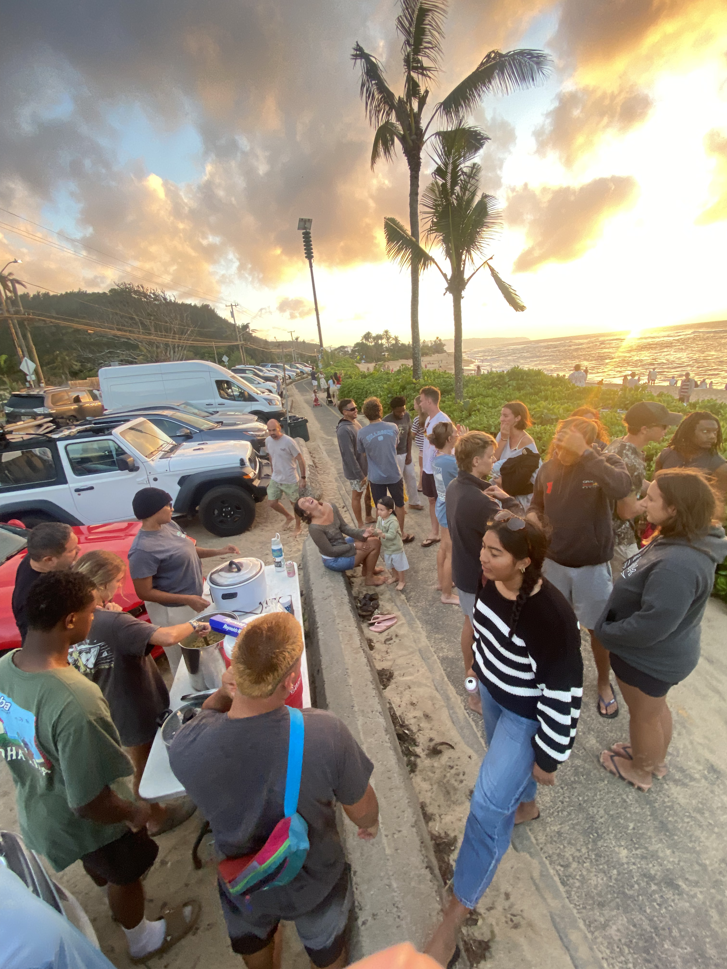 People gathered along a coastal road during sunset, some waiting in line at a food station, with parked cars and palm trees, ocean waves, and a cloudy sky in the background.