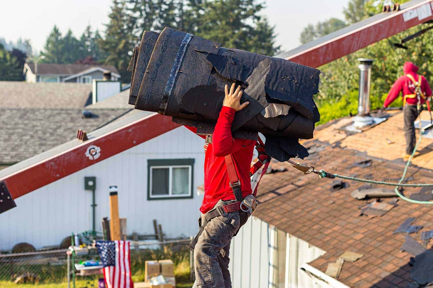 Worker on roof carrying a bundle of roof shingles, with another worker in the background working on the roof.