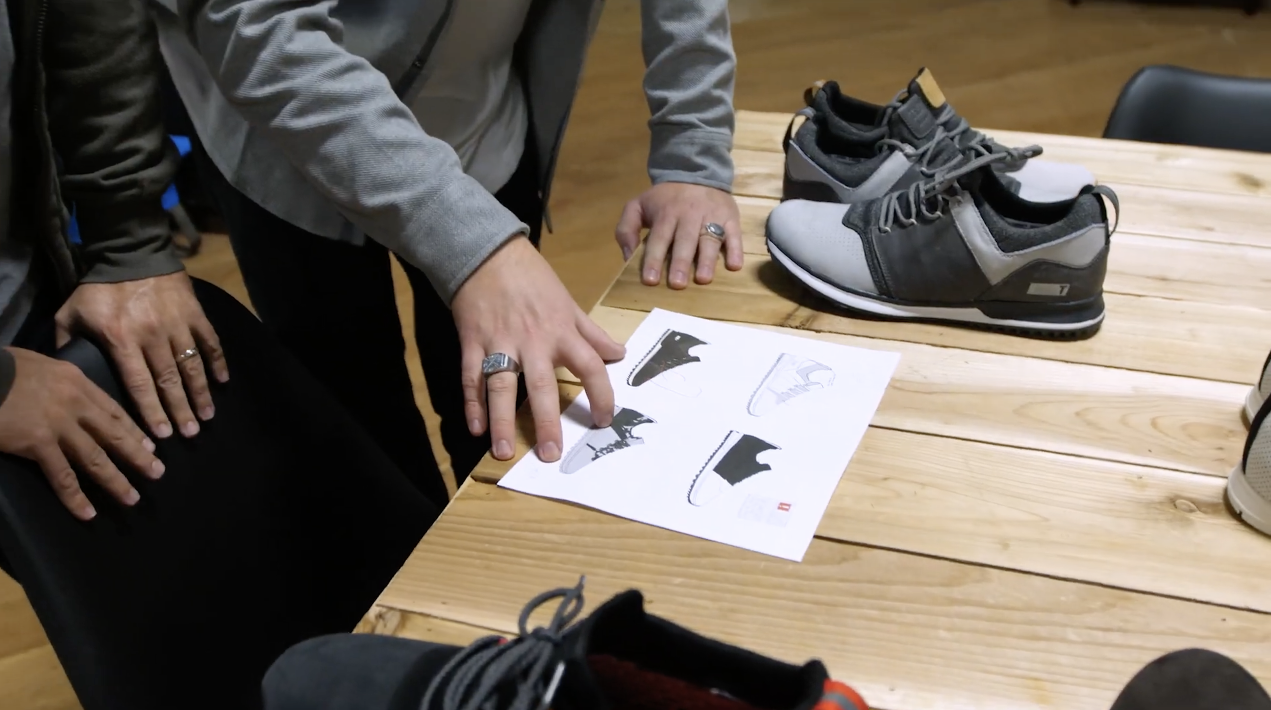 Person arranging gray and black sneakers on a wooden table, with shoe design sketches and an instructional sheet.