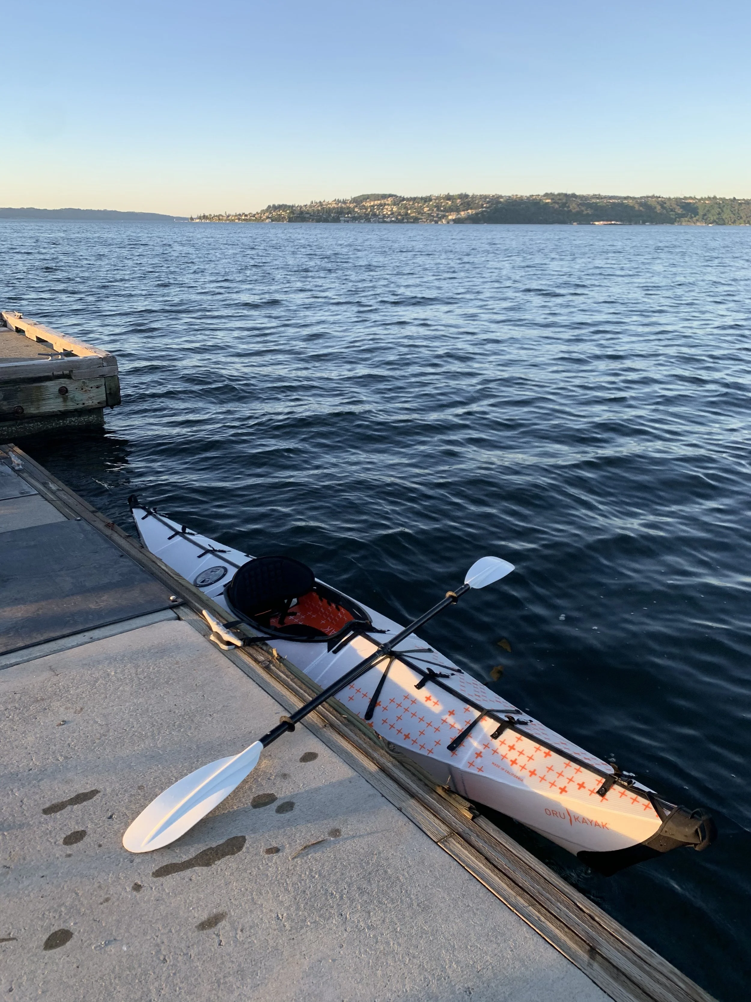 A white and orange kayak with two paddles resting on a concrete dock beside a body of water with a distant hillside in the background.