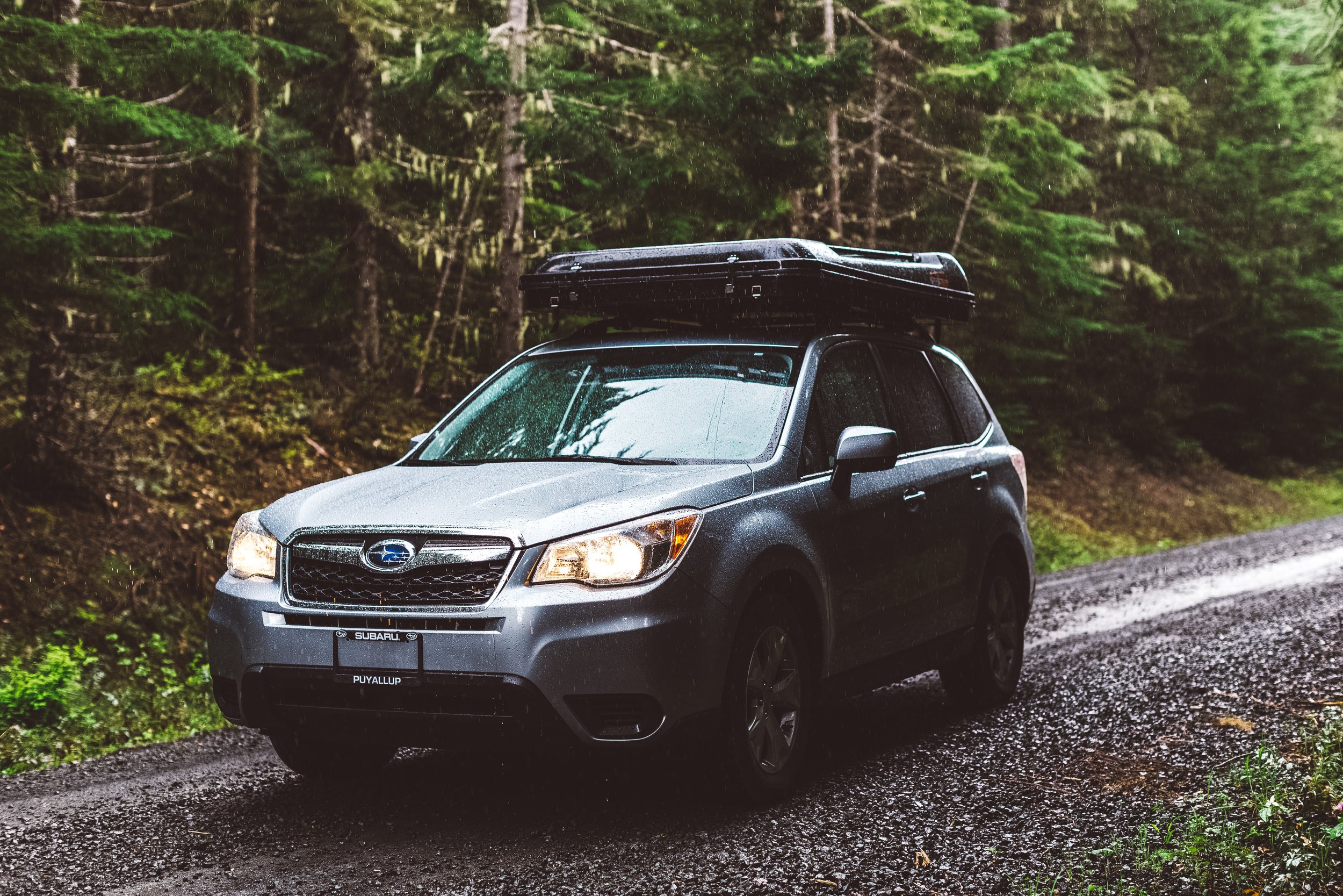 A silver Subaru SUV driving on a wet gravel road in a forested area, with trees and greenery in the background, during rainy weather.