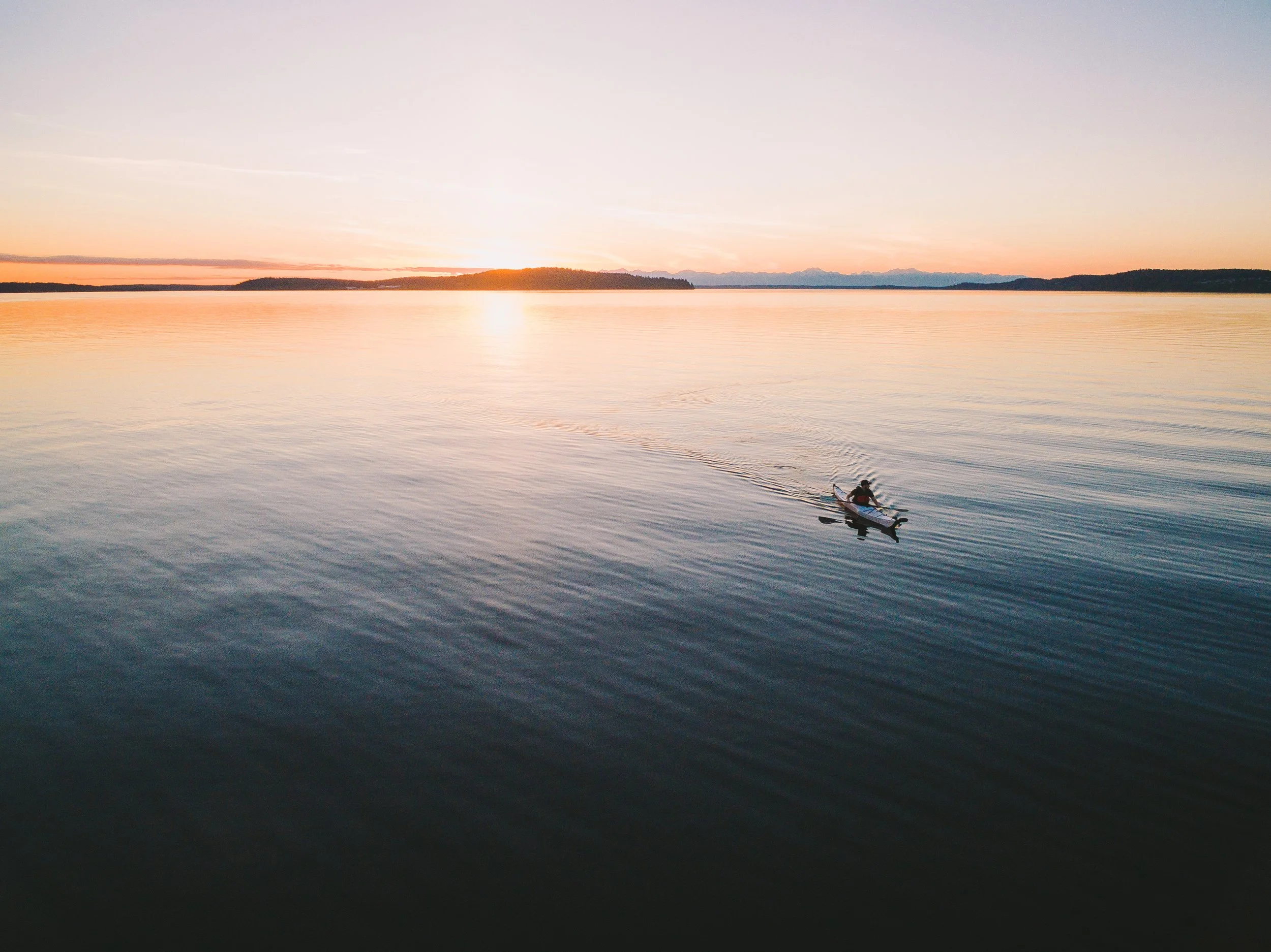 Sunset over a large body of water with a small boat and two people in it, with distant land and mountains in the background.