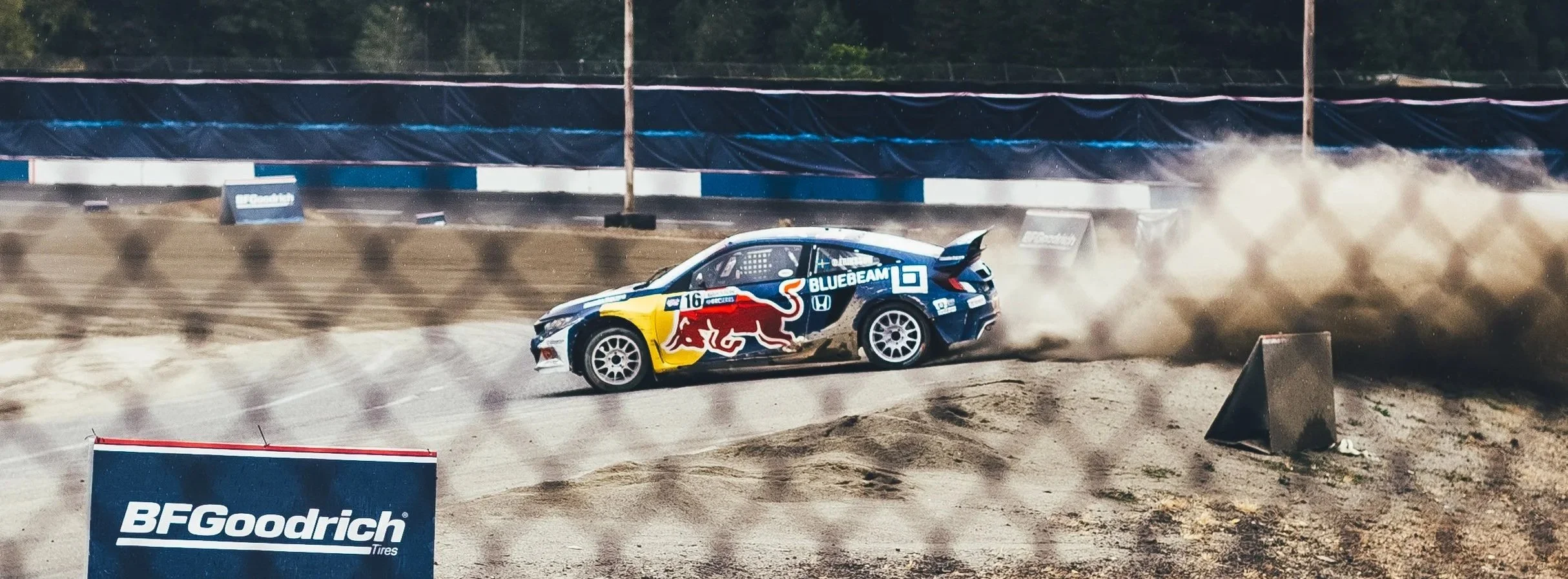 A rally car with Red Bull branding skids off the dirt track, creating a cloud of dust and debris, with a gravel barrier and sponsor signs in the background.