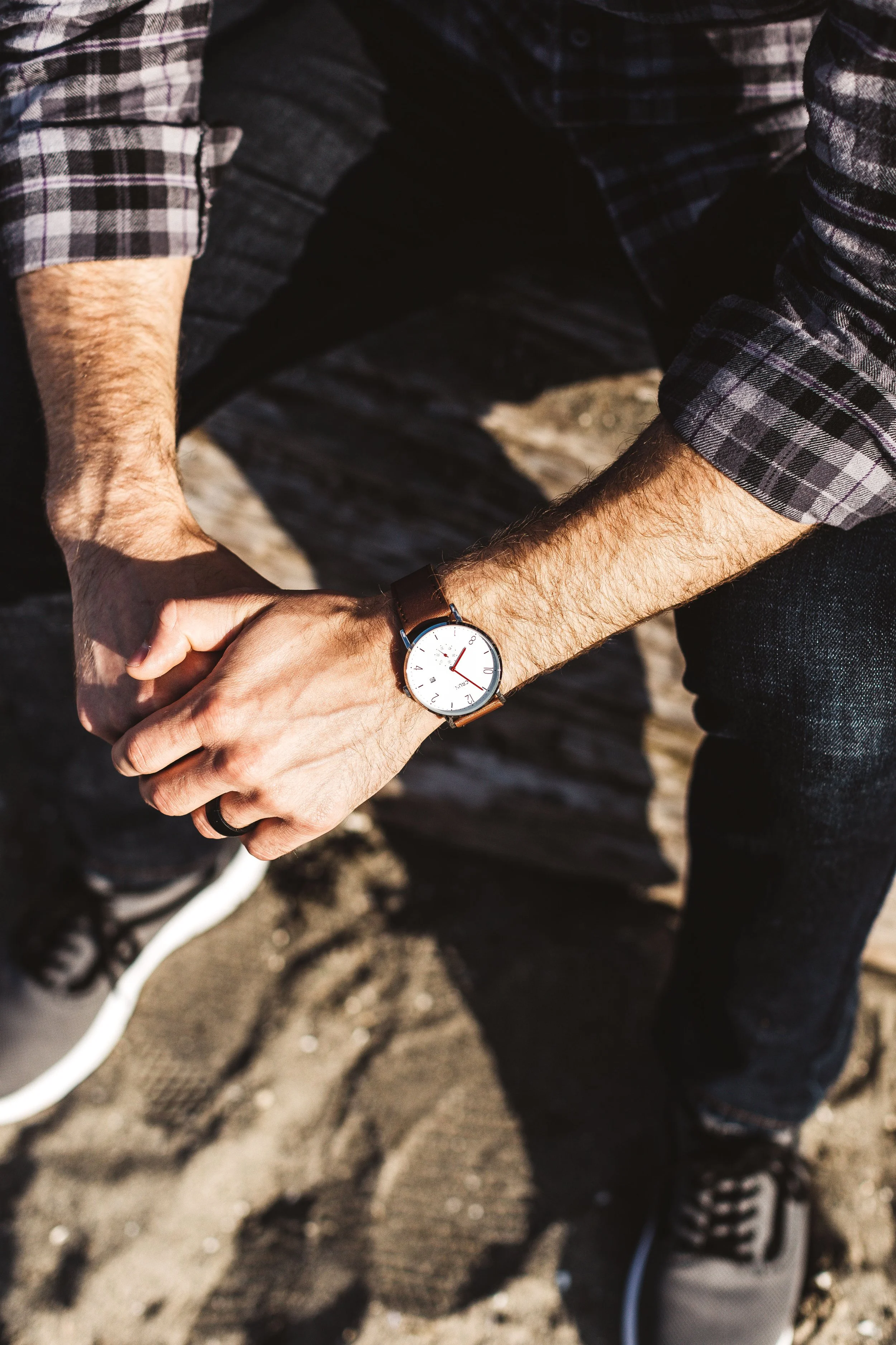 Two people holding hands outstretched, standing on a sandy surface, one wearing a plaid shirt with rolled-up sleeves, a wristwatch, and sneakers.