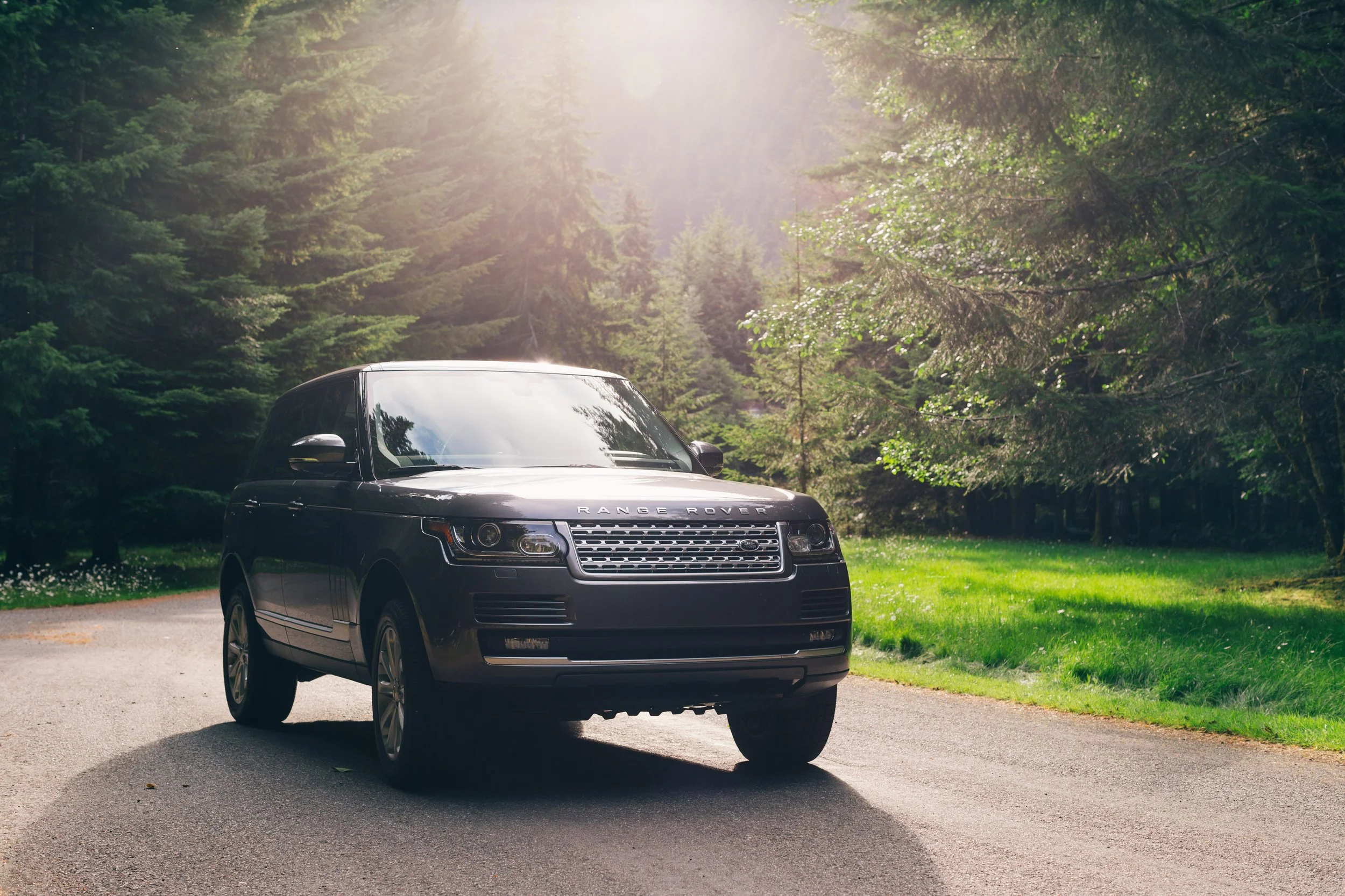 A black Range Rover parked on a paved driveway in a green wooded area with sunlight filtering through the trees.