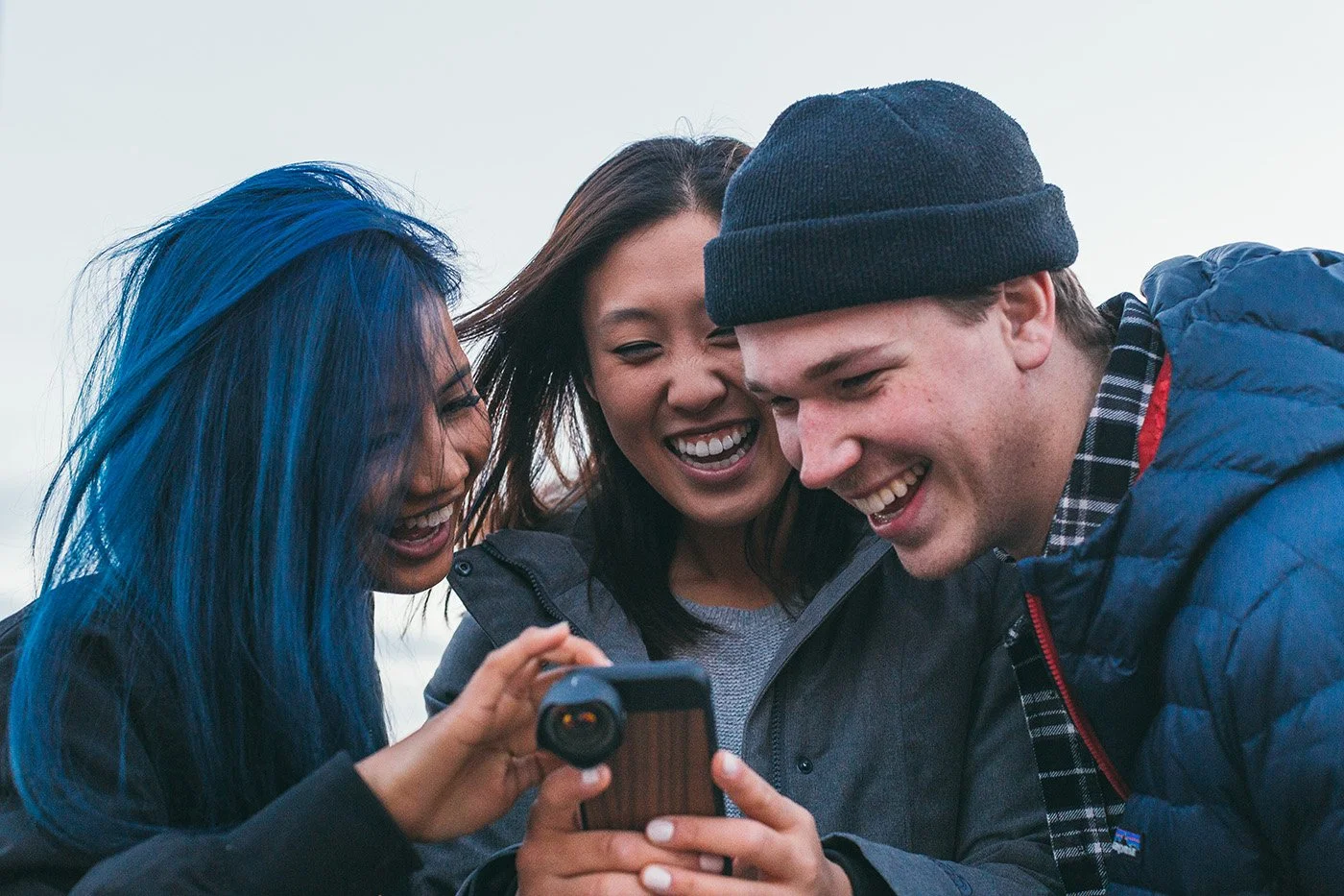 Three people smiling together while looking at a smartphone fitted with a Moment camera lens.