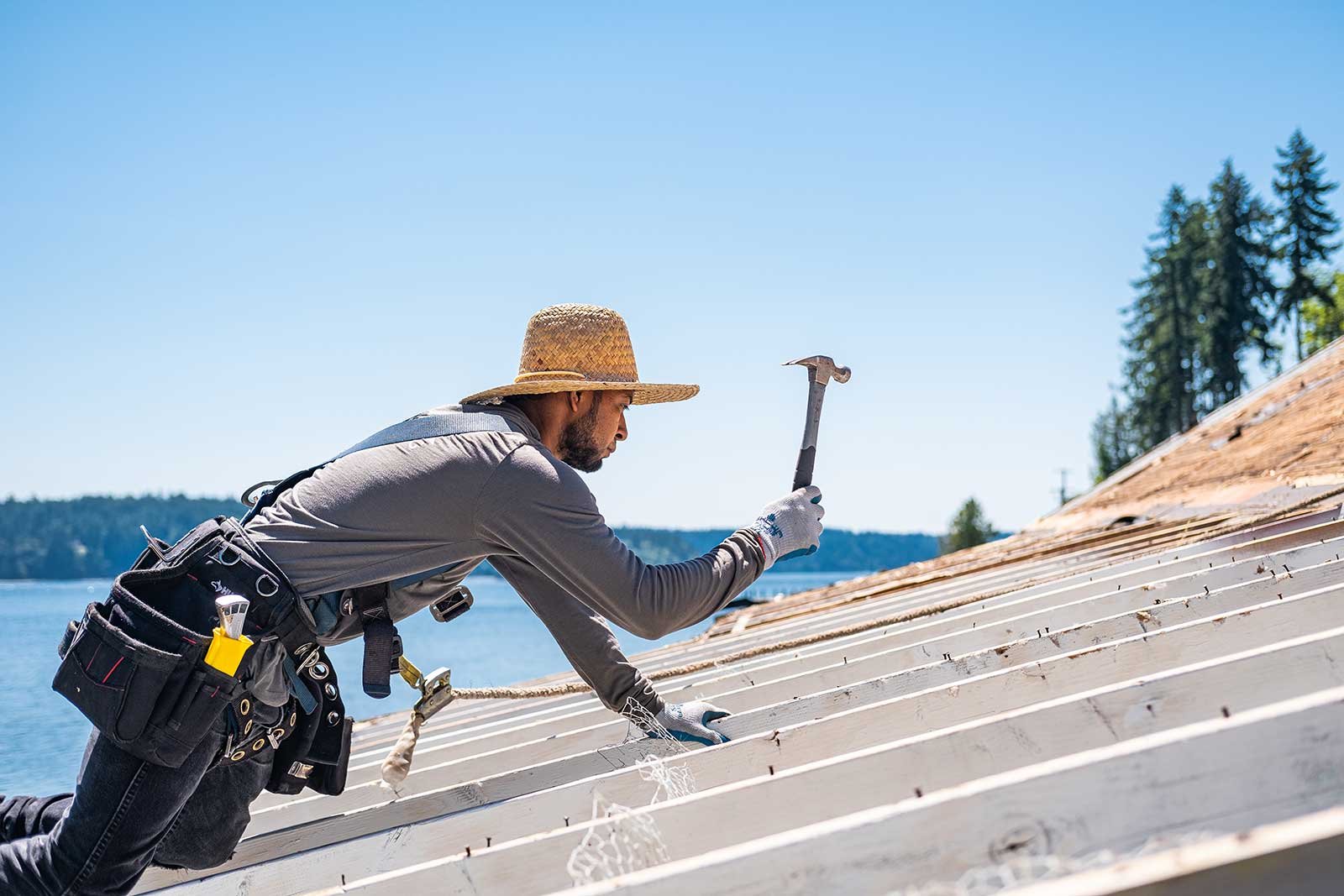 A man wearing a straw hat, gray long sleeve shirt, and work belt with tools kneels on a boat roof, using a hammer during a sunny day near a body of water, with trees and a clear sky in the background.