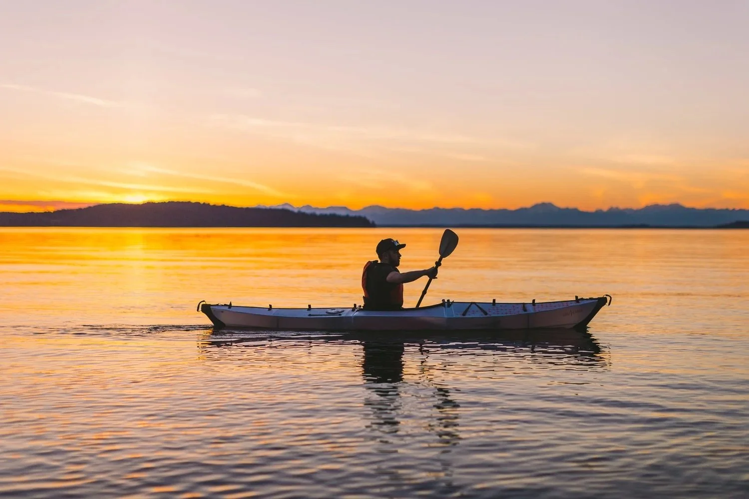 A person kayaking on a calm body of water during sunset, with a vibrant orange sky and a silhouette of distant mountains in the background.