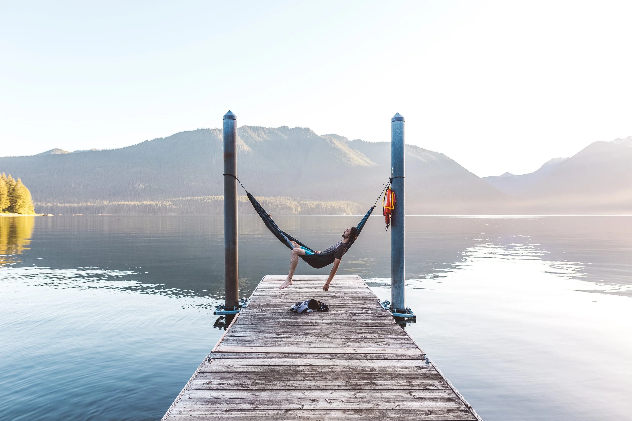 Person relaxing in a hammock on a wooden dock extending into a calm lake, with mountains in the background.