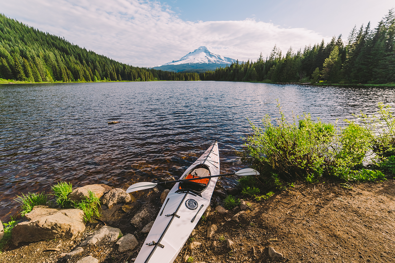 A kayak resting on the rocky shore of a lake with a mountain in the background and lush green trees lining the lake.