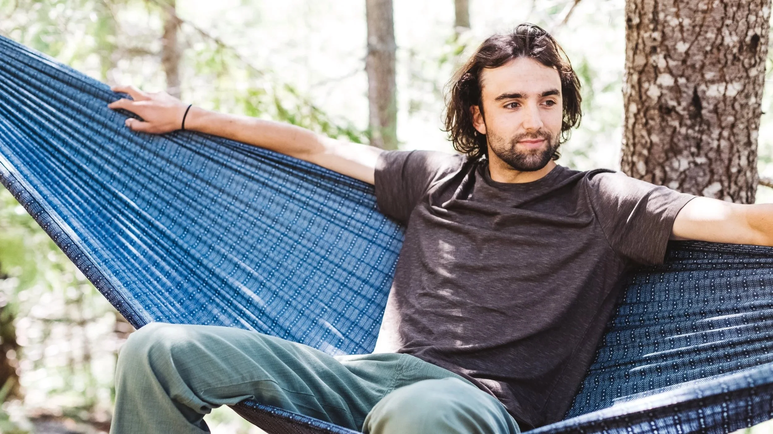 A young man with dark hair and beard relaxing in a blue hammock in a forest setting.