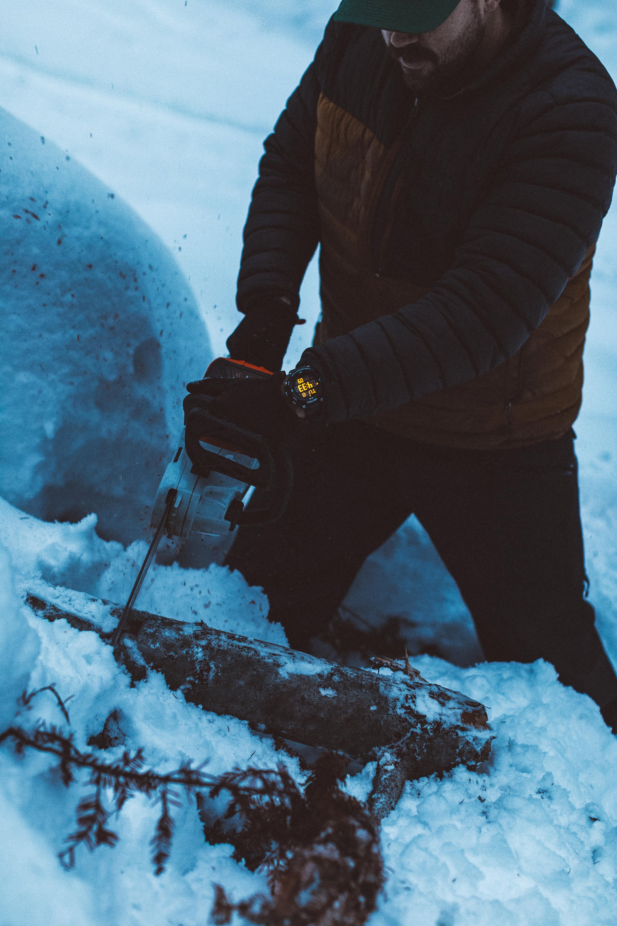 Person kneeling in snow, cutting a fallen log with a chainsaw
