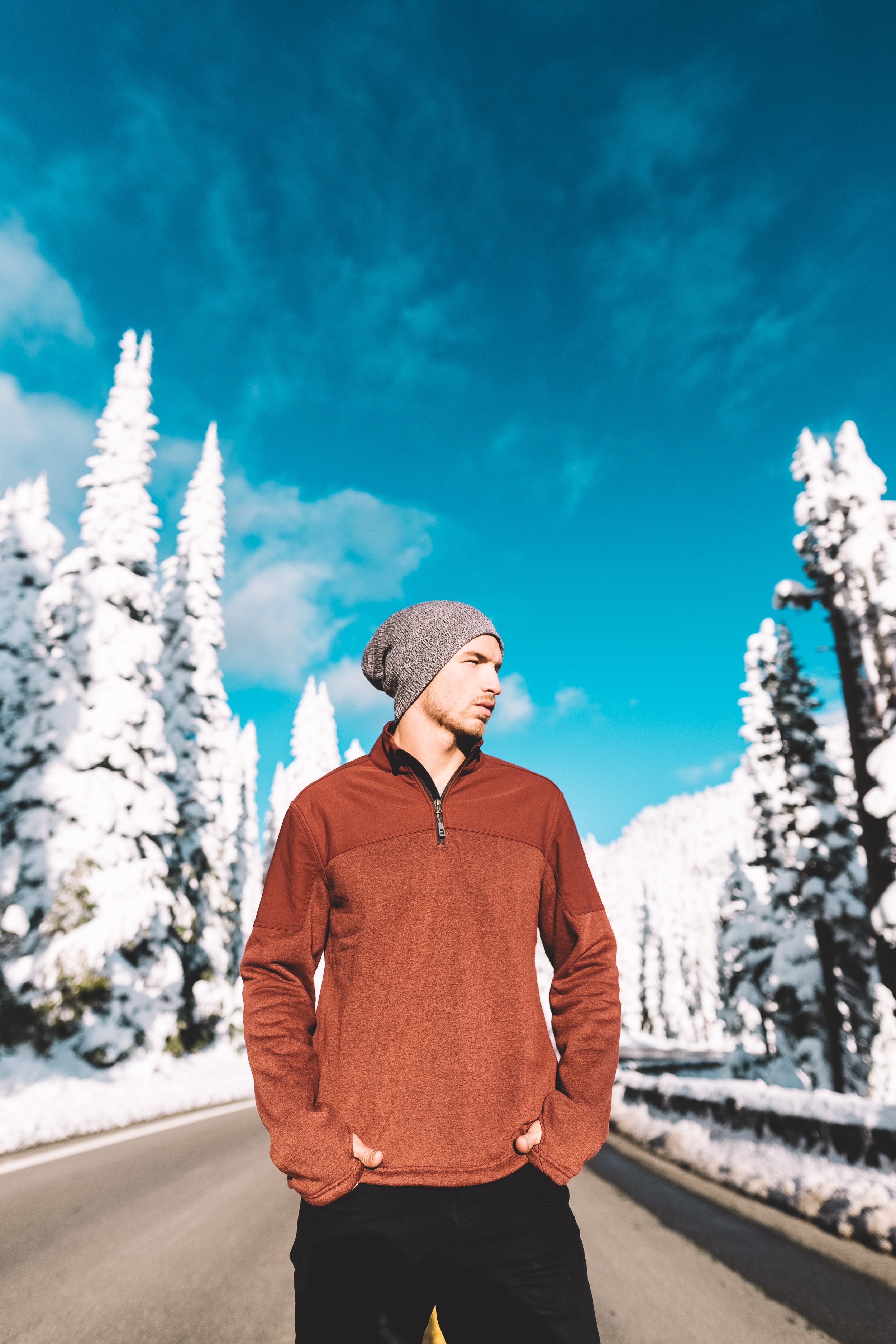 A man wearing a gray beanie and a brown jacket walking along a snowy mountain road with snow-covered trees against a blue sky.