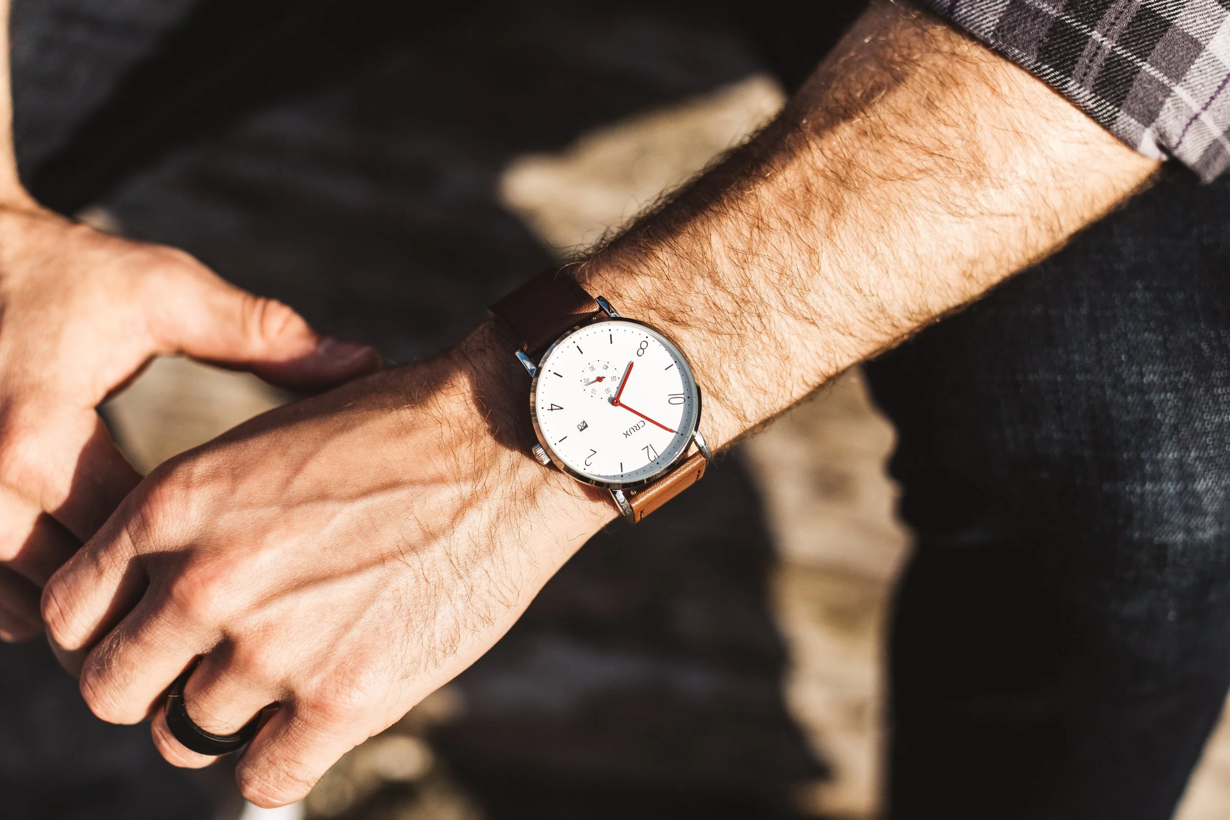 A person wearing a wristwatch with a white face and brown leather strap, checking the time outdoors with sunlight illuminating their arm.