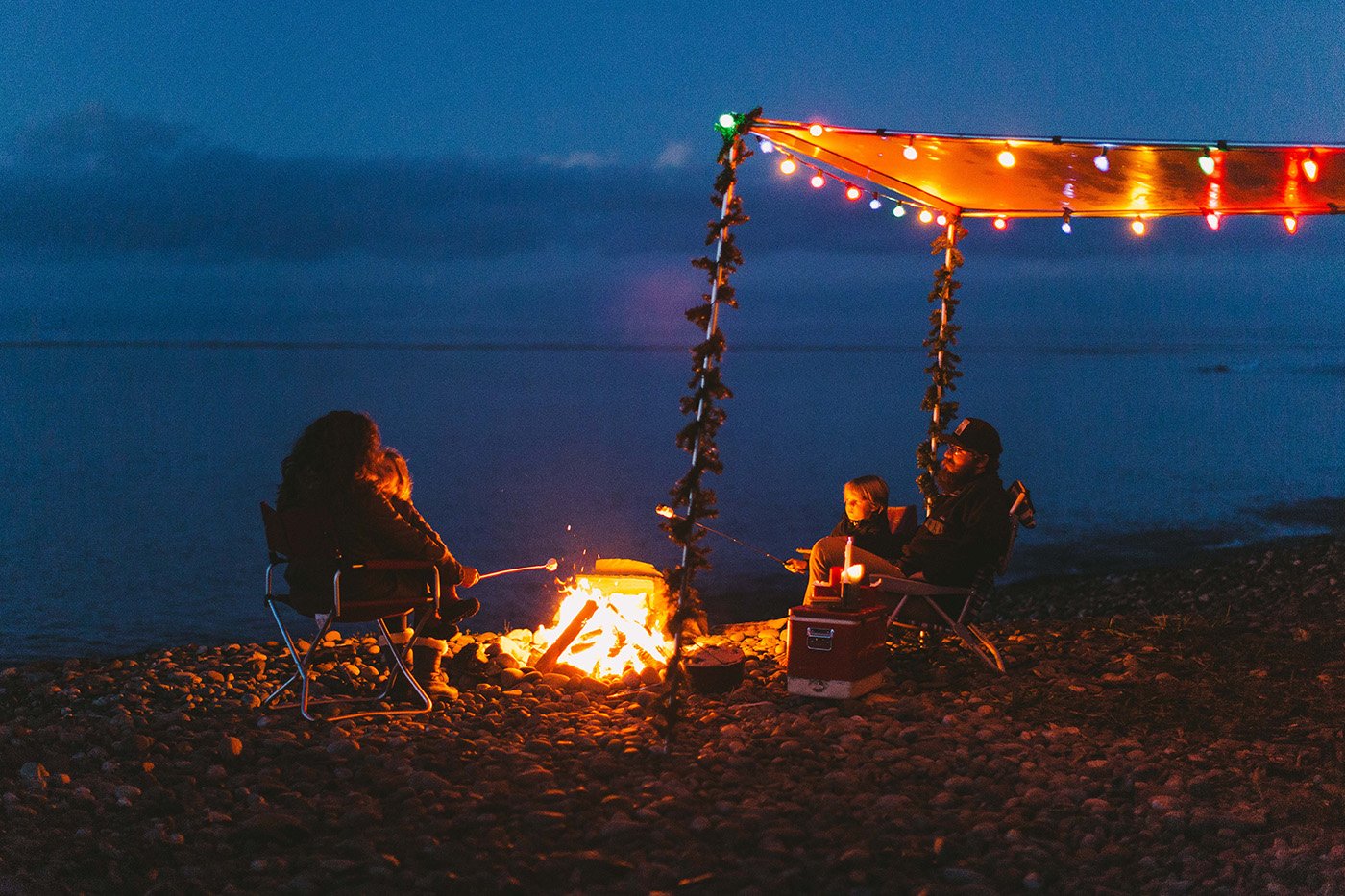 Friends gathered around a campfire beneath string lights on a beach at dusk.