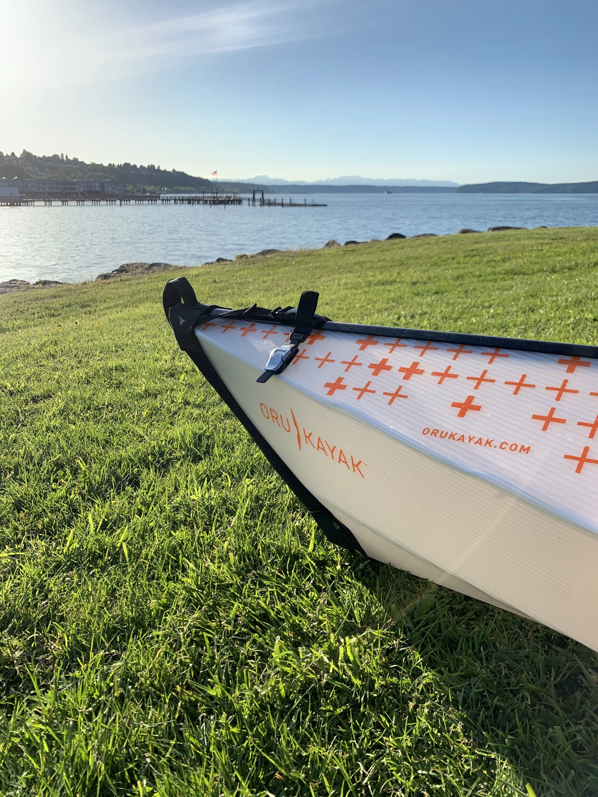 A kayak resting on green grass by a body of water, with a pier and distant hills under a clear blue sky.