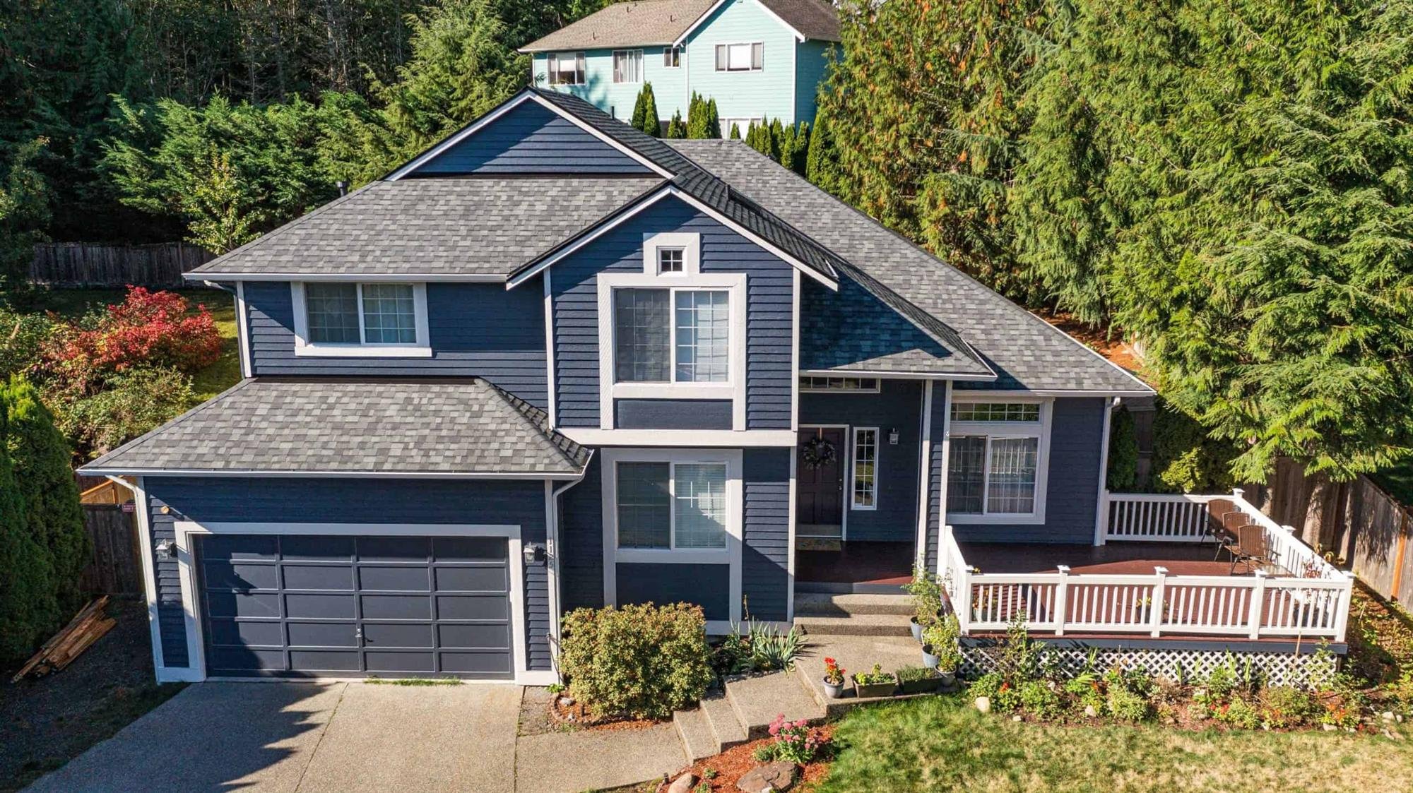 A two-story blue house with a gray roof, surrounded by green trees and a white fence. The house has a garage, multiple windows, and a small deck with chairs.