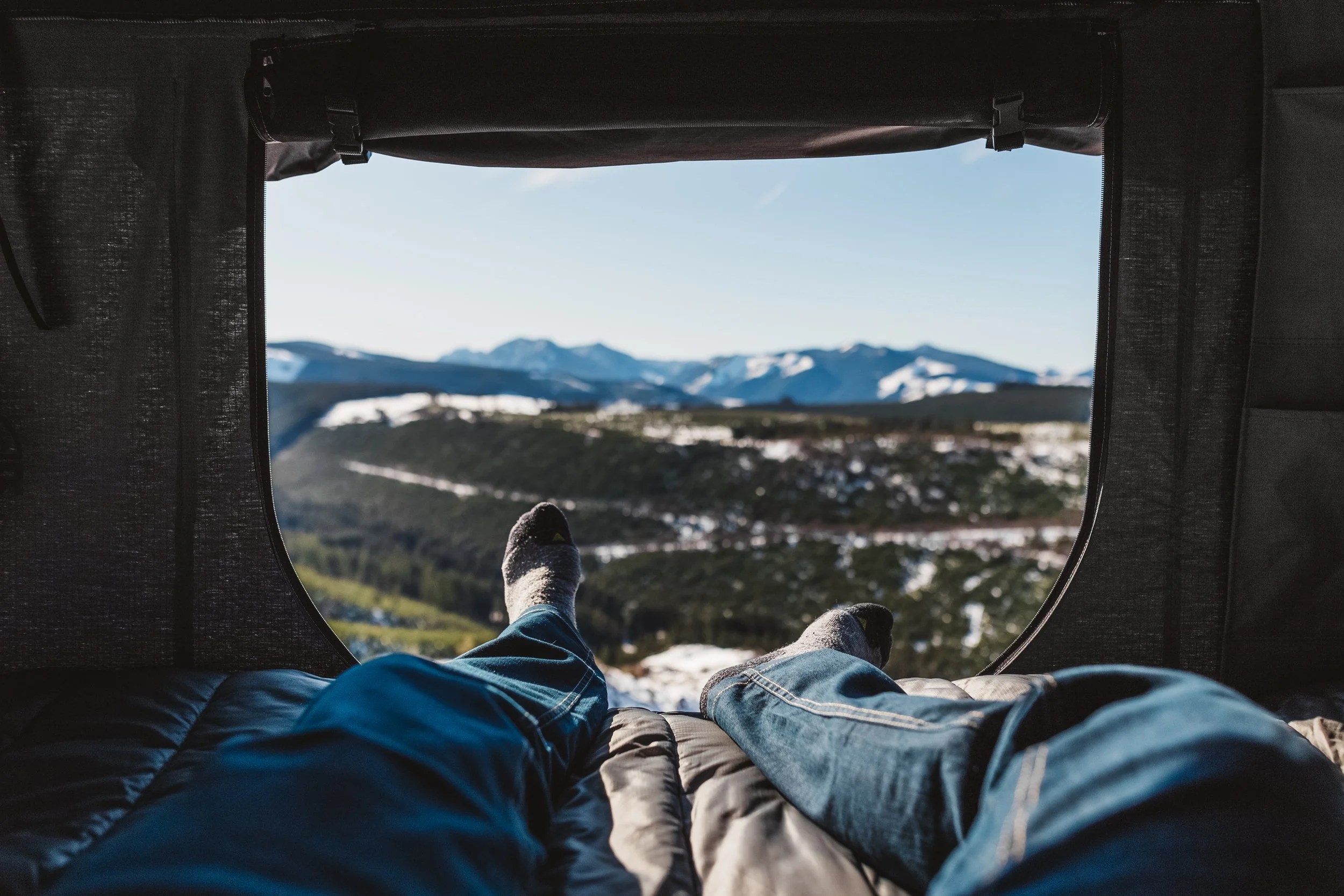 Inside a tent, two people are lying down and relaxing while looking out at a mountain landscape with snow-capped peaks and a clear blue sky.