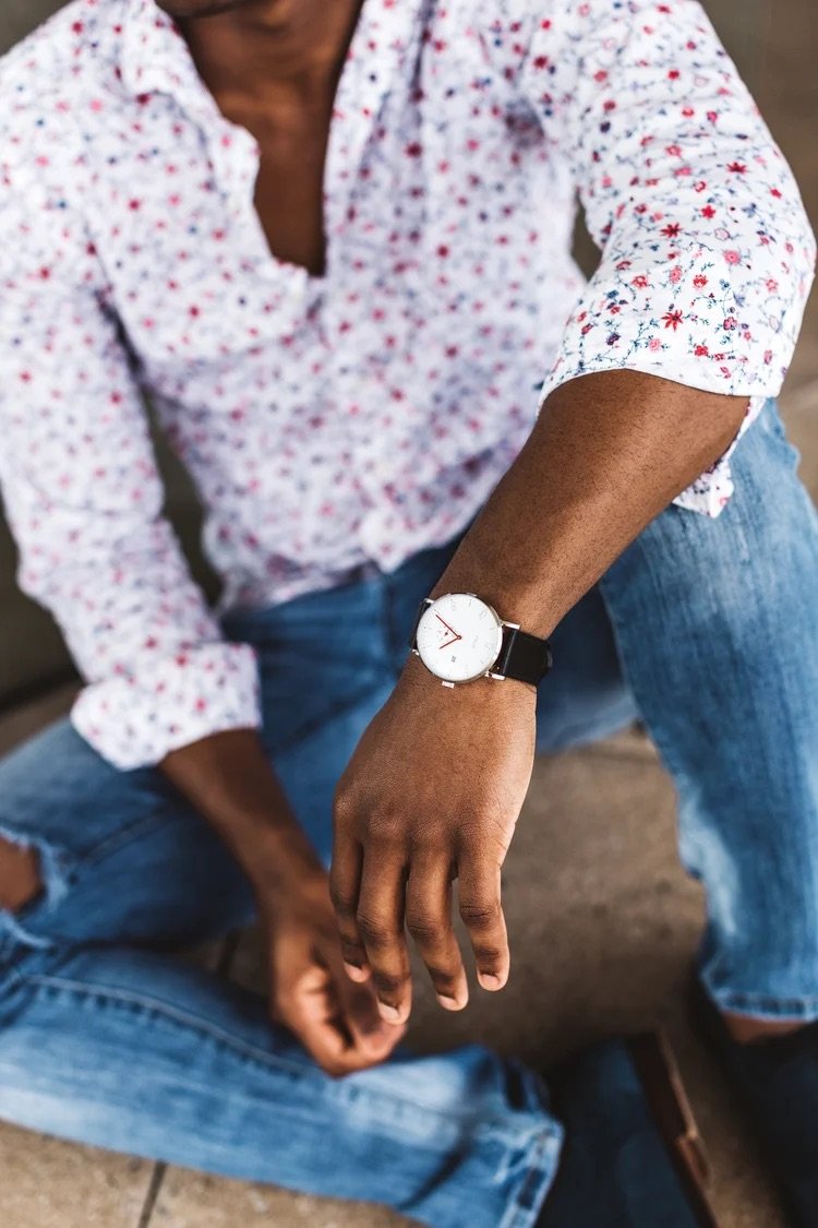 A person wearing a white shirt with a small red and blue floral pattern, blue jeans, and a wristwatch with a white face and black strap, sitting with legs crossed and leaning forward.