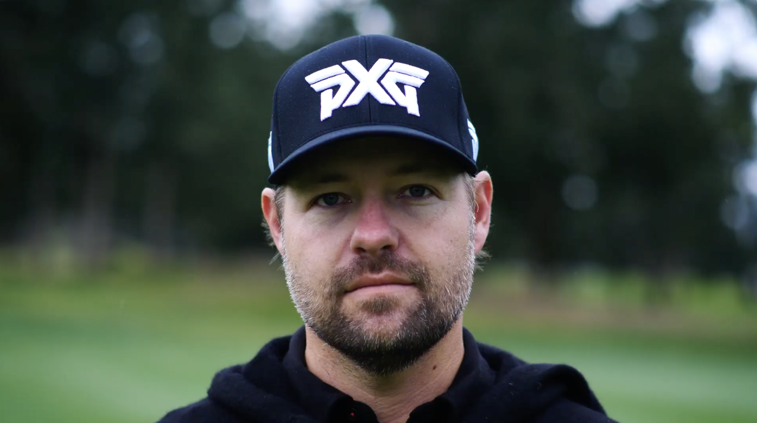 A man with a beard and blue eyes wearing a black cap with white logo and a black shirt, outdoors with blurred greenery in the background.