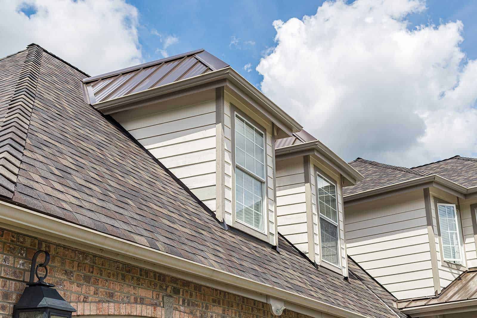 Close-up of a residential house's roof with brick and beige siding, featuring two large windows and a small metal roof section with clouds in the sky.