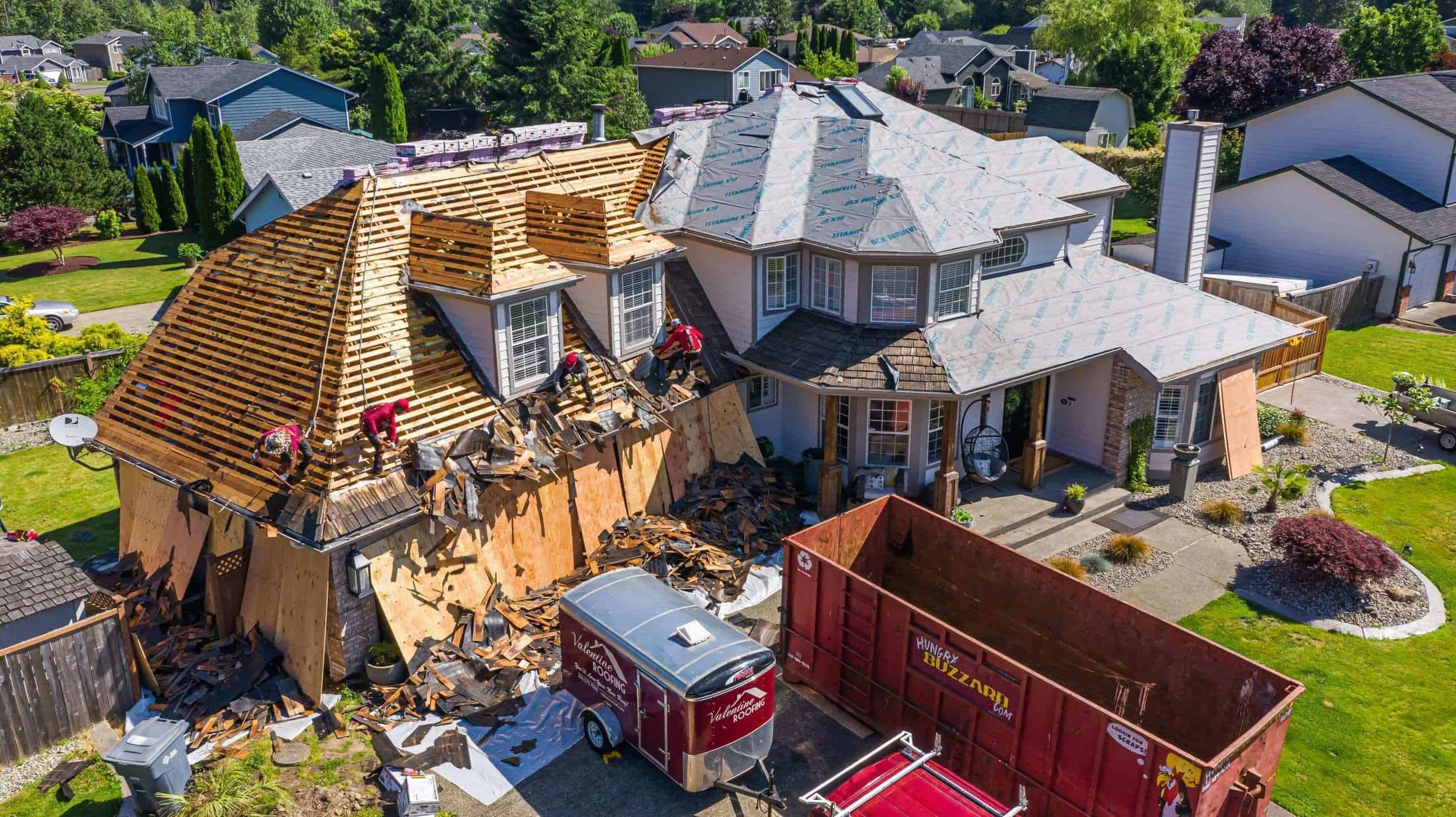 An aerial view of a house under construction with workers tearing off the roof, with a dumpster and cars parked nearby, in a neighborhood with green lawns and trees.