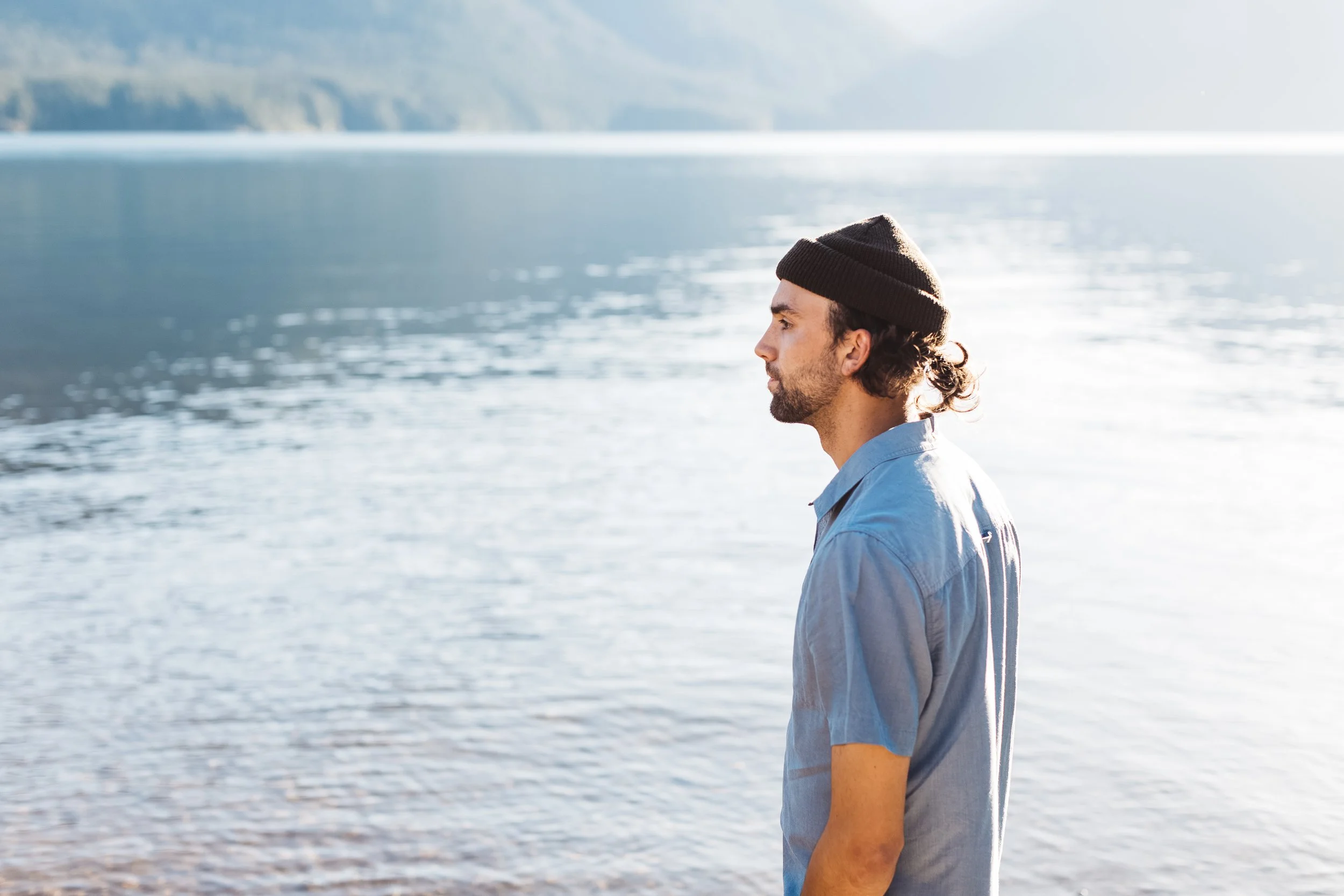 A man wearing a brown beanie and a light blue shirt standing by a body of water with mountains in the background.
