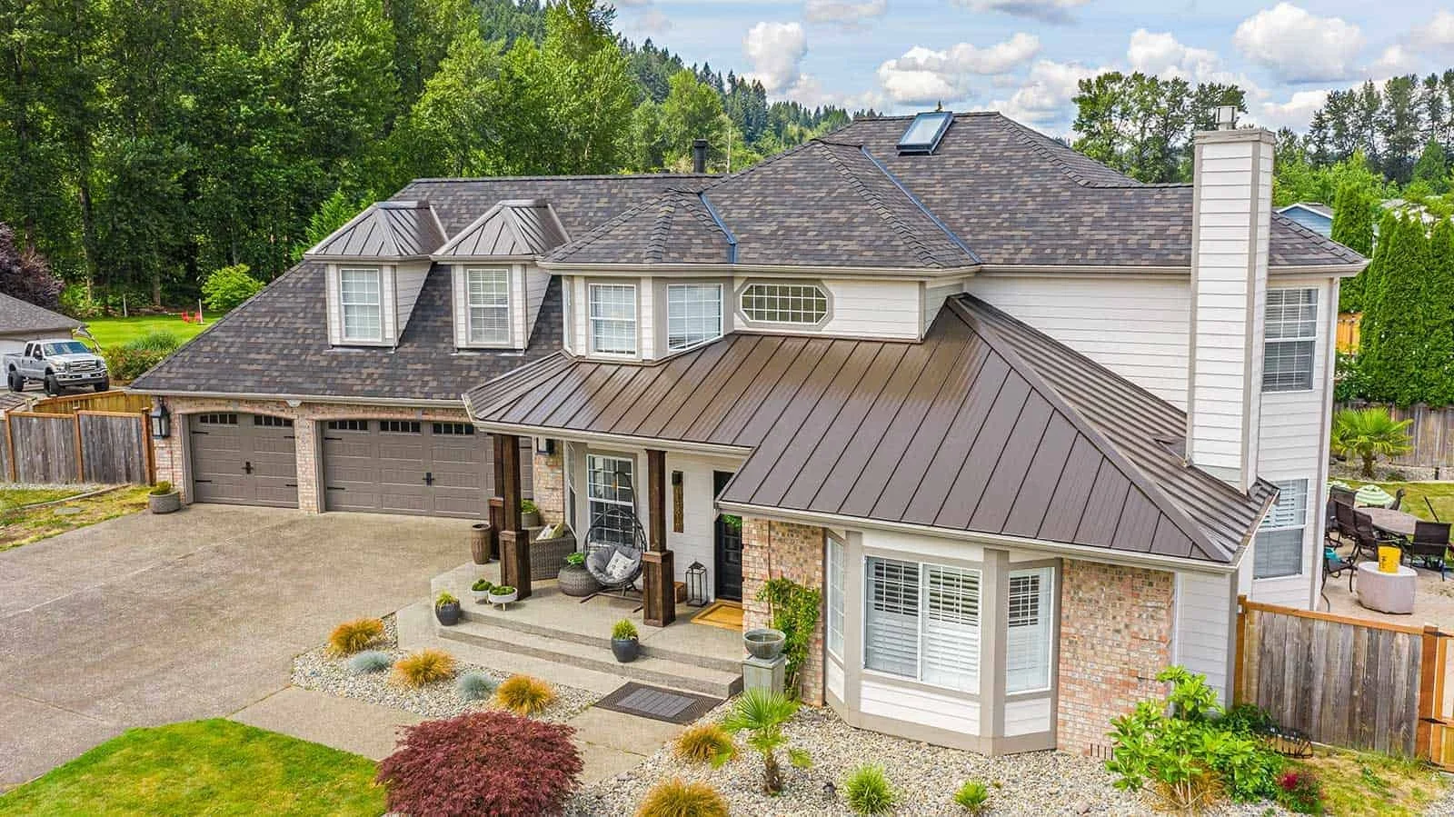 A large two-story house with a dark shingled roof, multiple windows, and a small front porch with outdoor seating. The yard features a concrete driveway, landscaped garden beds, and a wooden fence, with green trees and cloudy sky in the background.