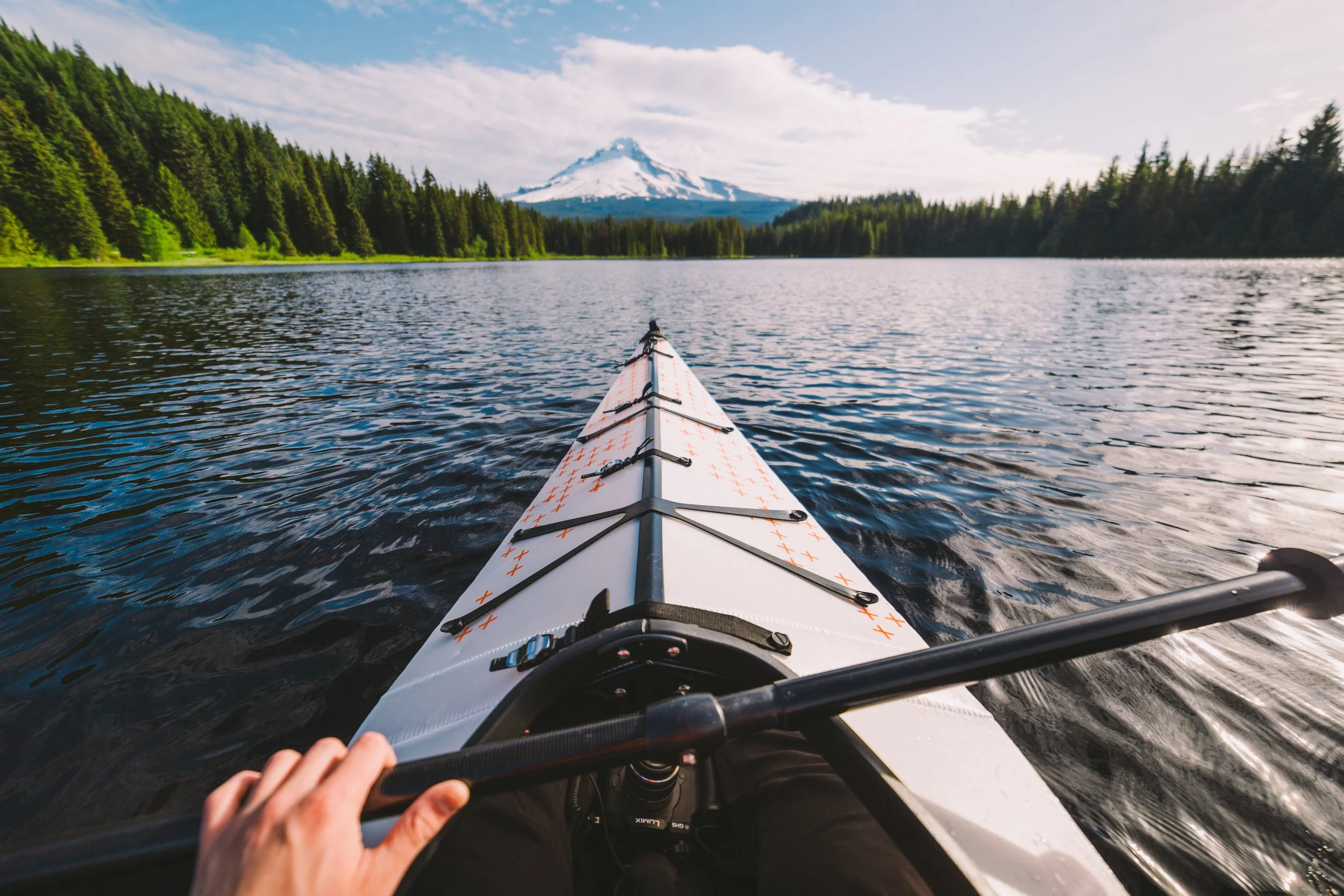 A person kayaking on a lake with a snow-capped mountain and green forest in the background.