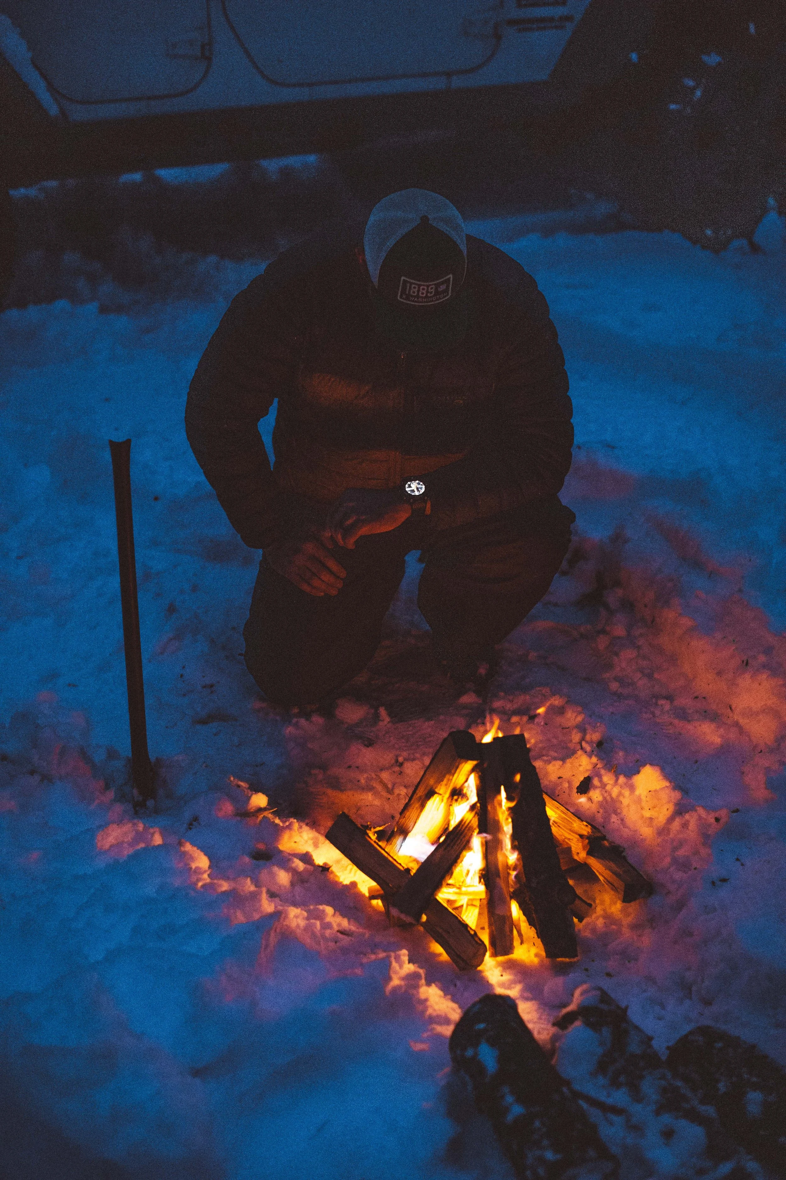 Person in winter clothing is kneeling in snow around a small campfire at dusk or night, with a brick structure in the background.