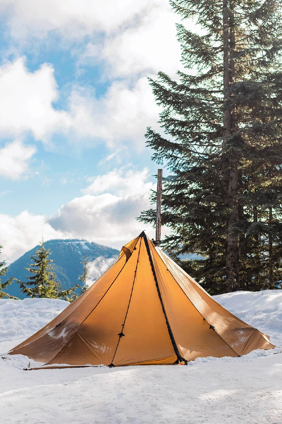 Tan canvas tent pitched among snow-covered mountains and evergreen trees.