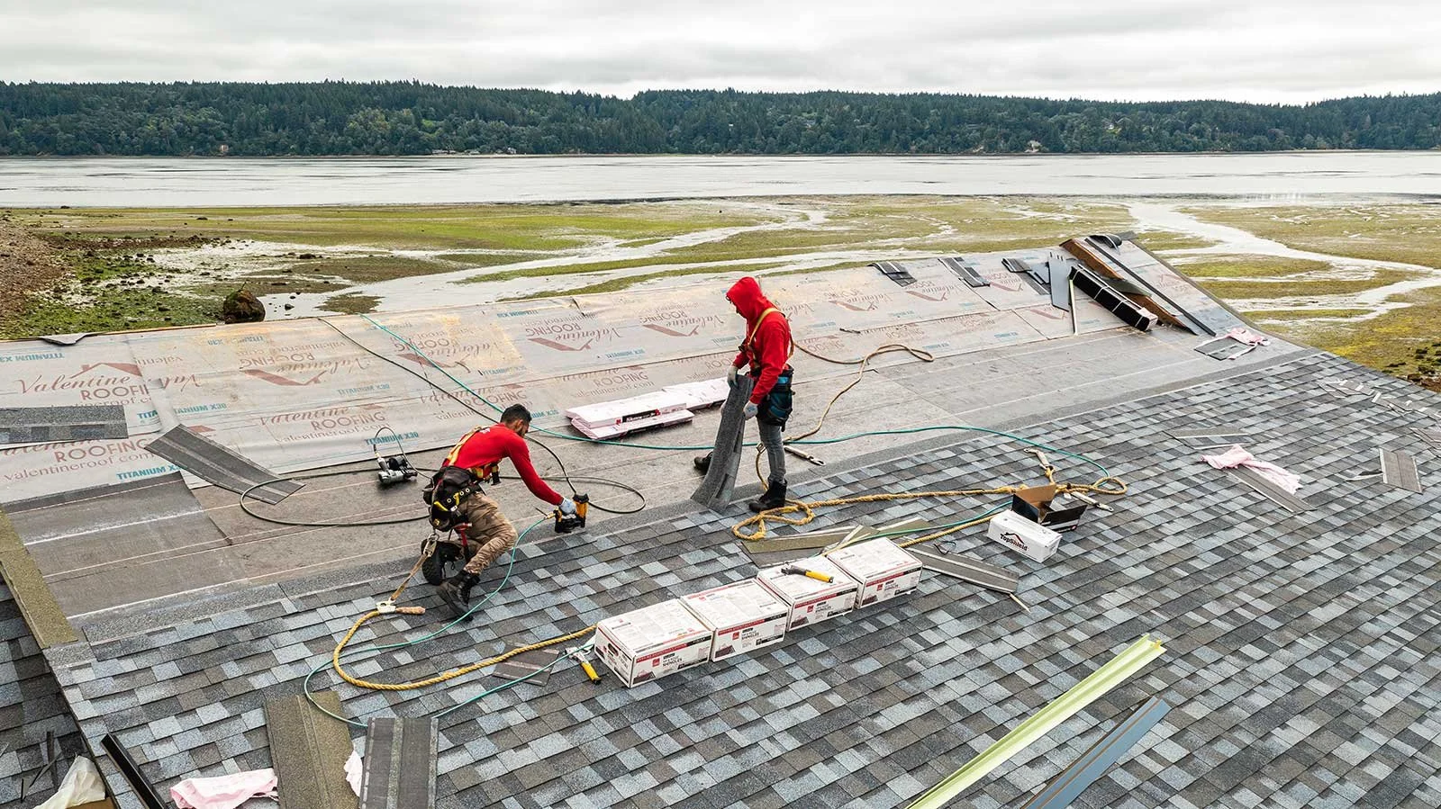 Two workers installing roofing shingles on a large sloped roof near a body of water and a green forest, with cloudy skies overhead.