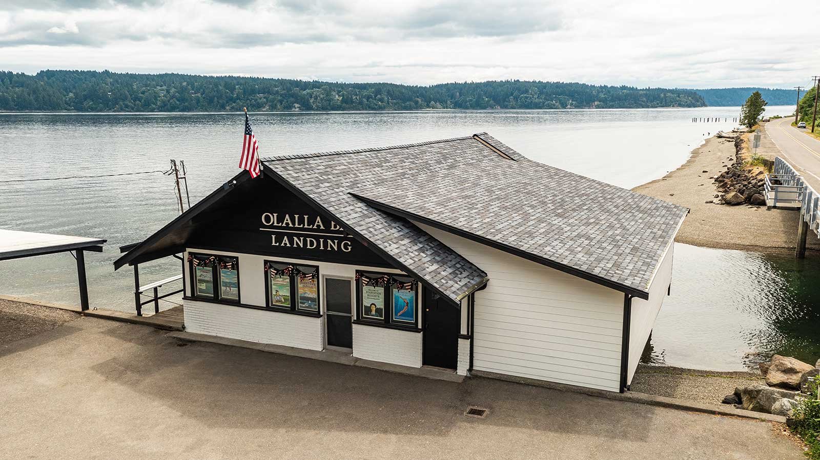A building on the shoreline labeled 'OLALLA BAY LANDING' with an American flag on top, overlooking a body of water with a tree-lined shore in the background.