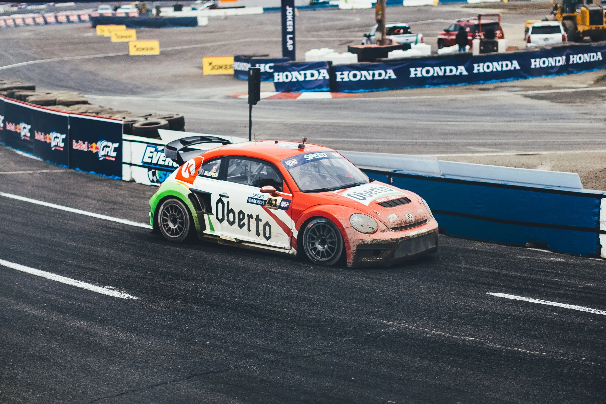 A race car with a red, white, and green color scheme driving on a race track, with tire barriers and branding banners in the background.
