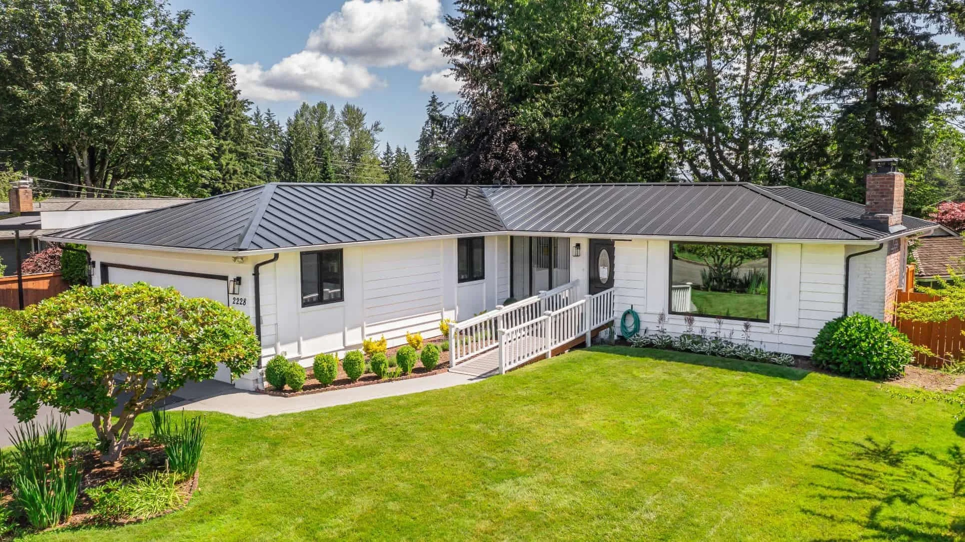 A white single-story house with a black metal roof, a front porch with a white railing, and well-maintained green lawn and landscaping. Tall trees surround the house, and a bright blue sky with scattered clouds is above.
