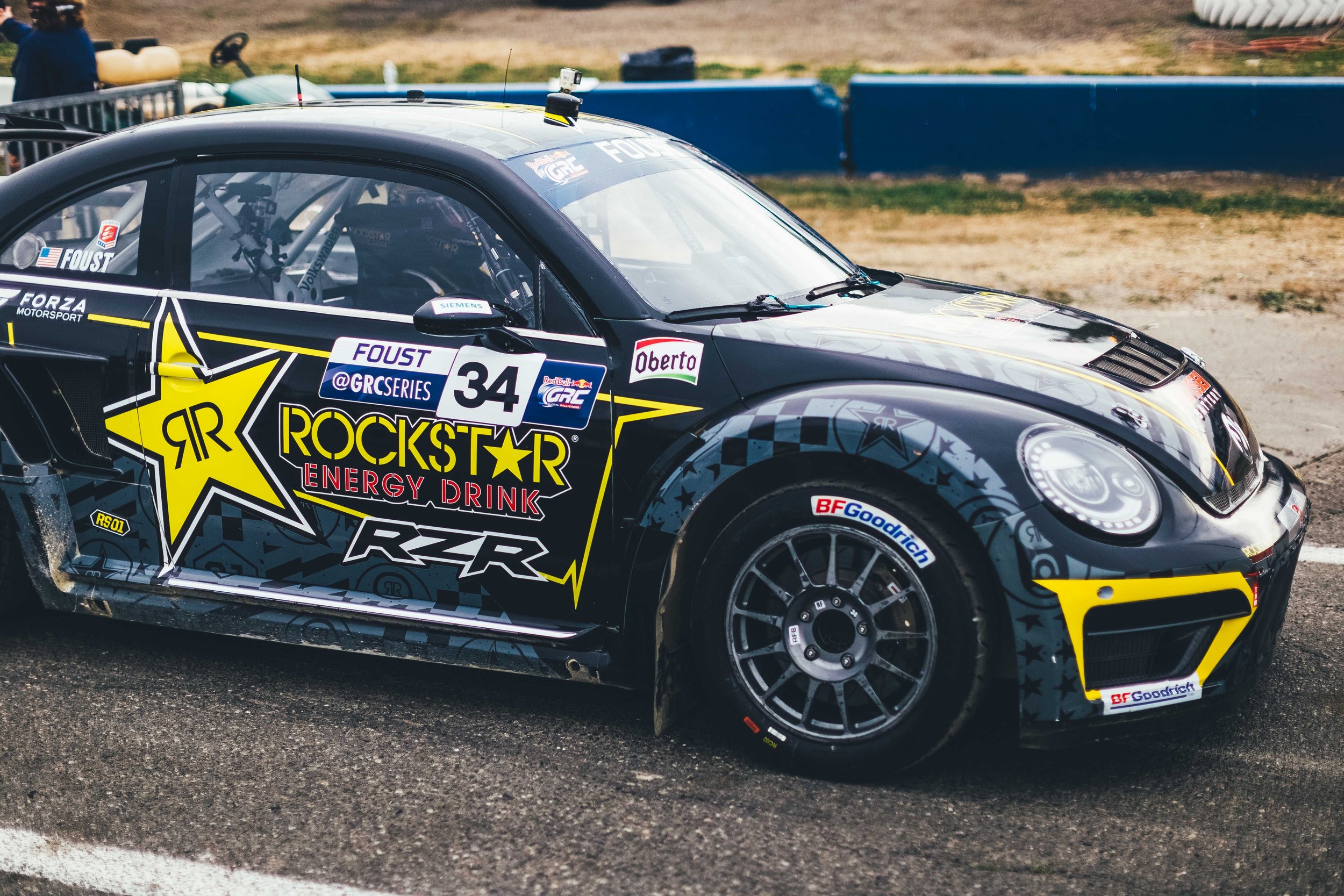 Close-up view of a racing car with Rockstar Energy Drink branding on the side, number 34, at a race track. The car is black with yellow accents, and features sponsor logos including Red Bull, Siemens, and BF Goodrich.