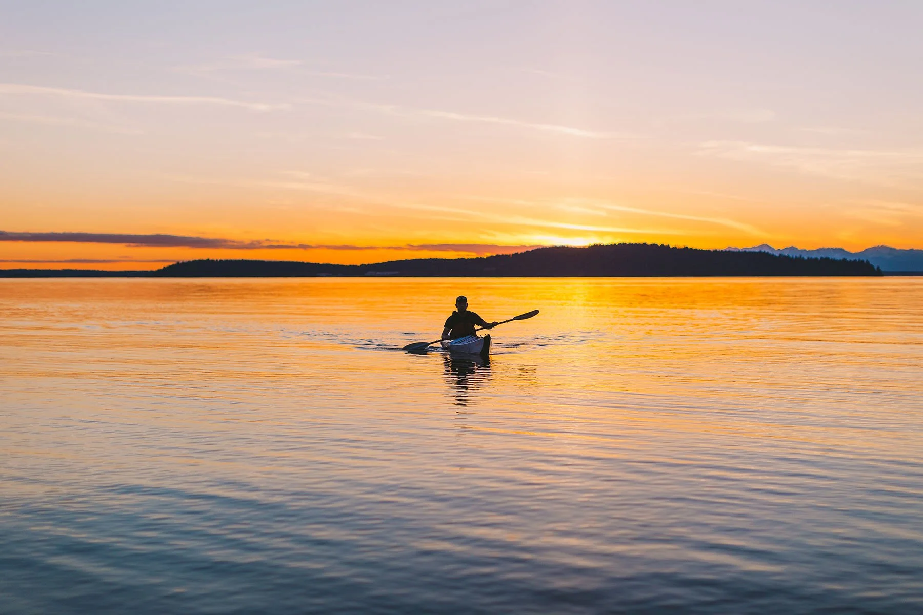 Kayaker paddling an Oru Kayak Bay+ across calm water at sunset.