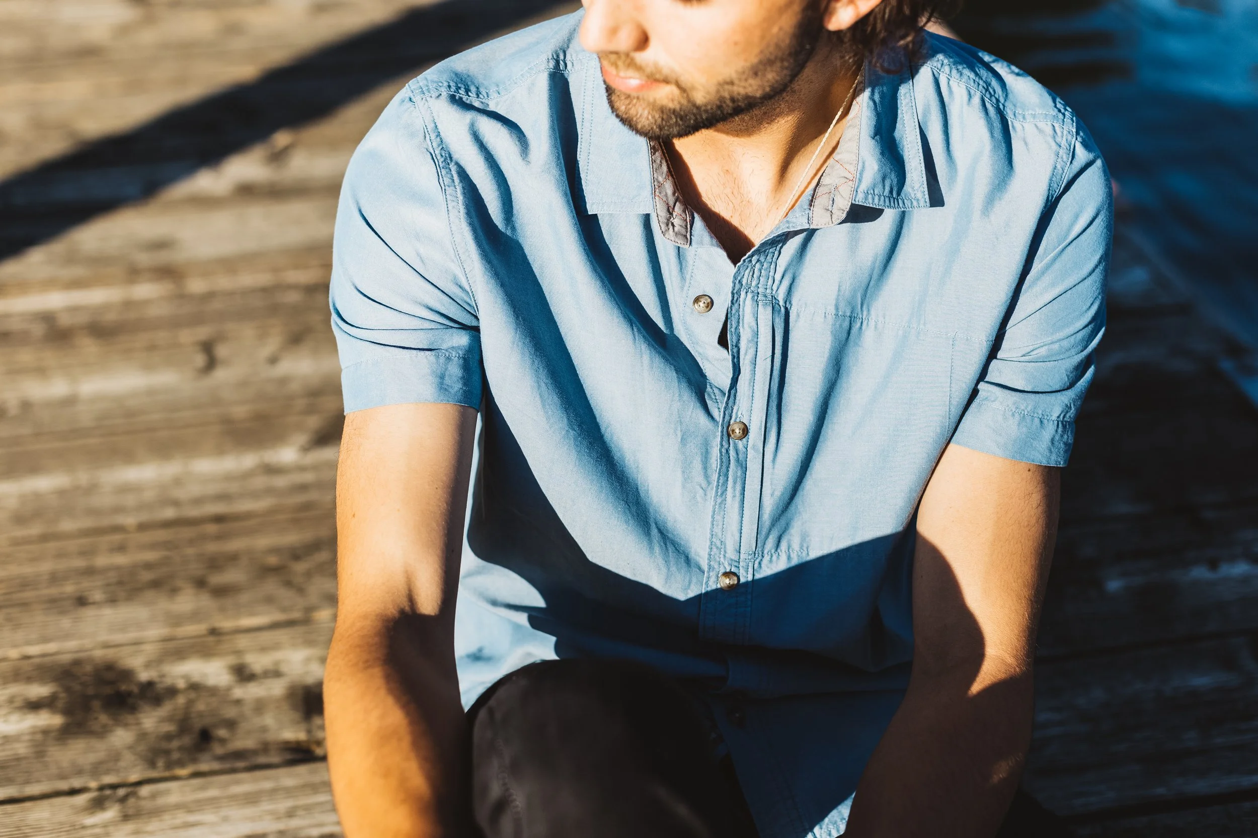 A young man with a beard wearing a light blue button-up shirt, sitting outdoors on a wooden surface, with water in the background, in sunlight.