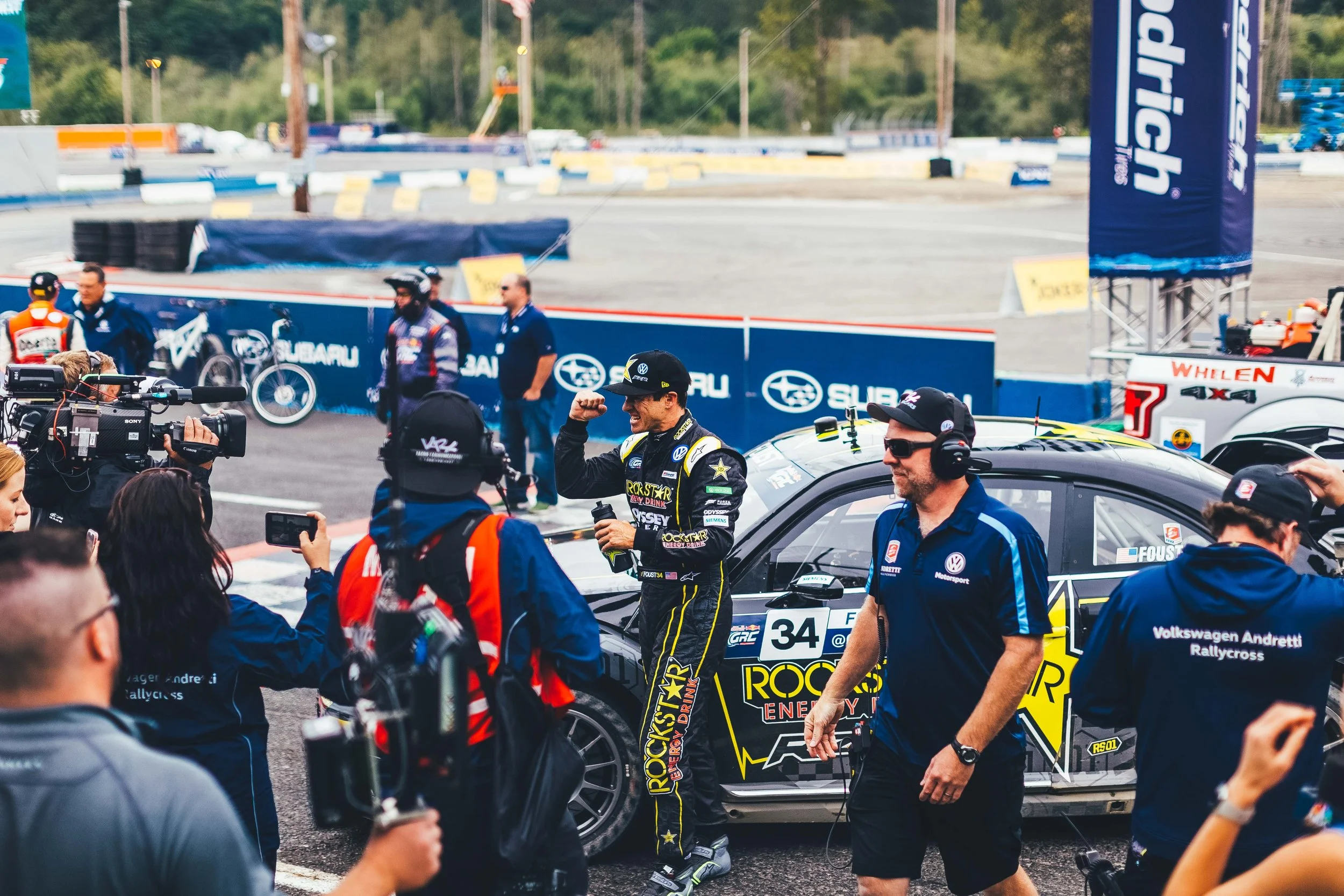 Race car driver celebrating in front of a race car at a race track, surrounded by team members and media.