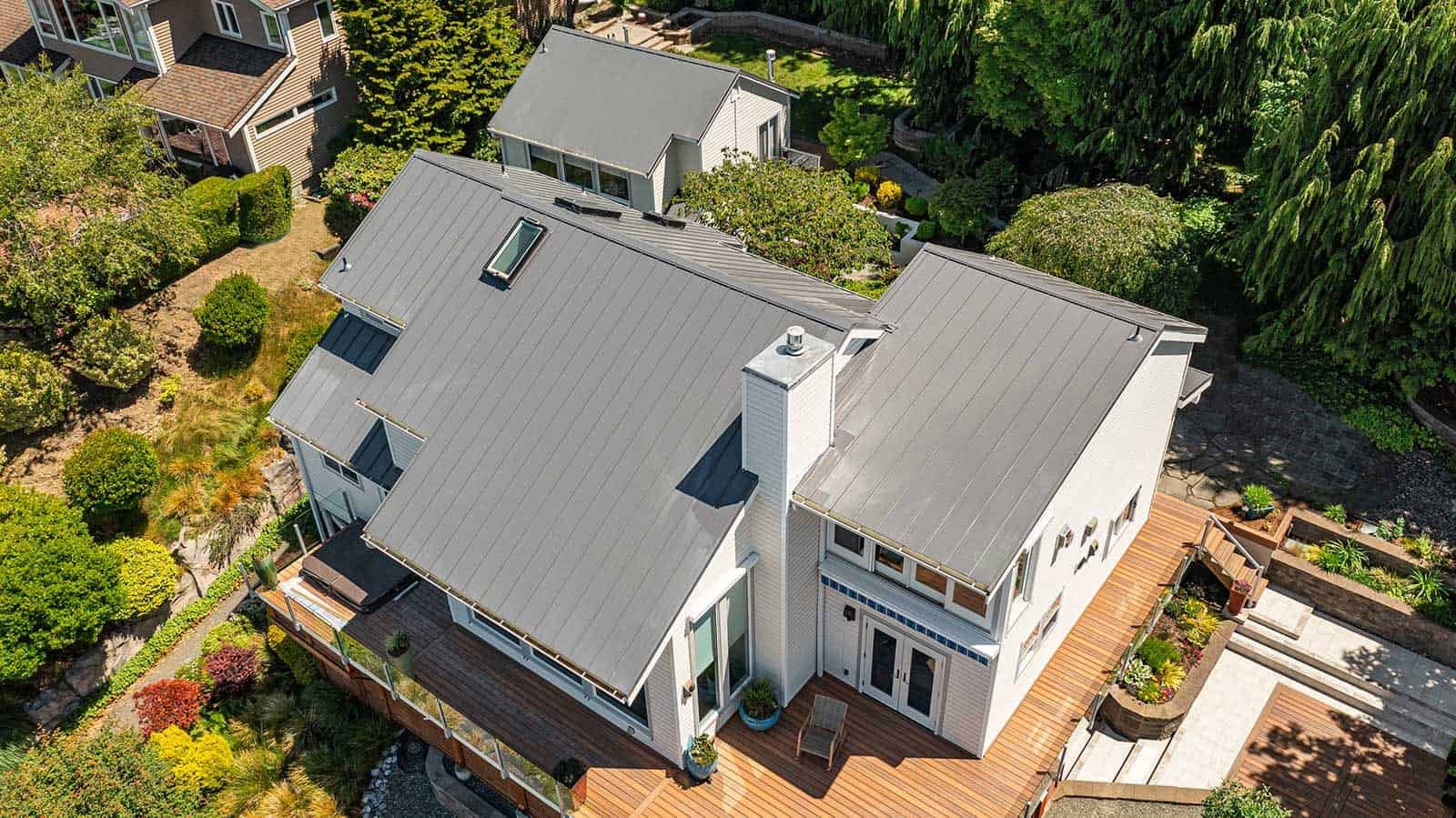 Aerial view of a modern two-story house with a metal roof, surrounding landscaped yard with trees, shrubs, and a wooden deck patio.