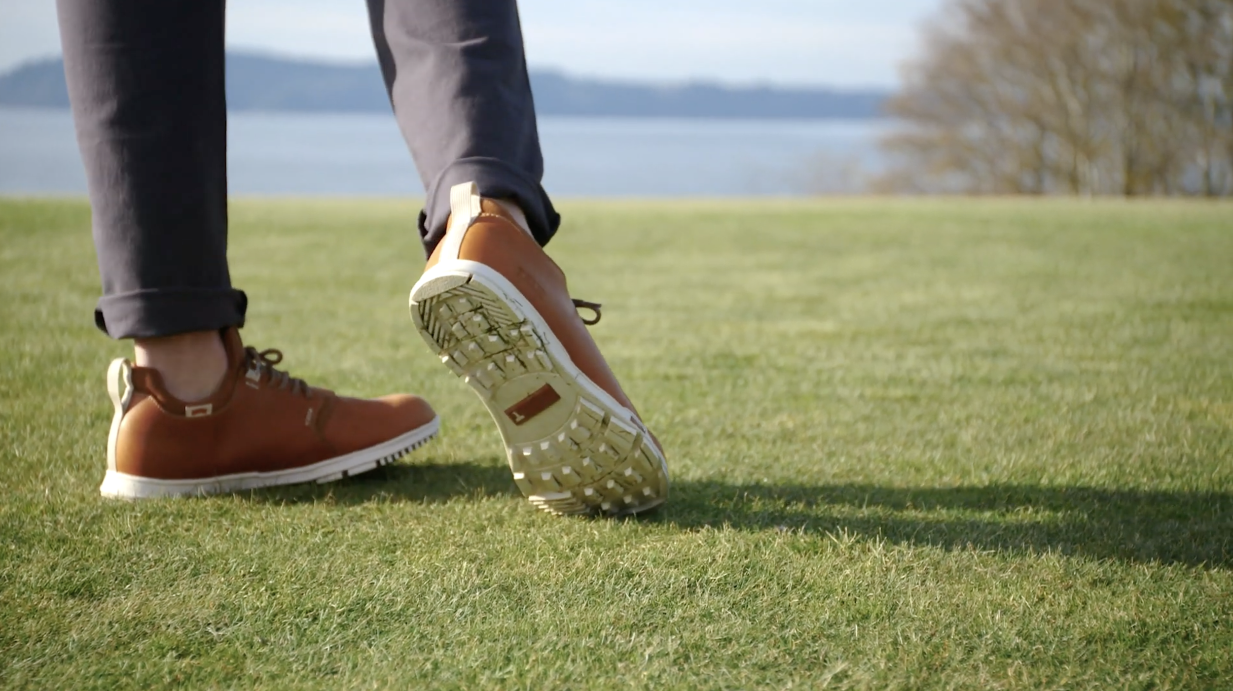 Close-up of person walking on grass, wearing brown boots and gray pants, with water and trees in the background.