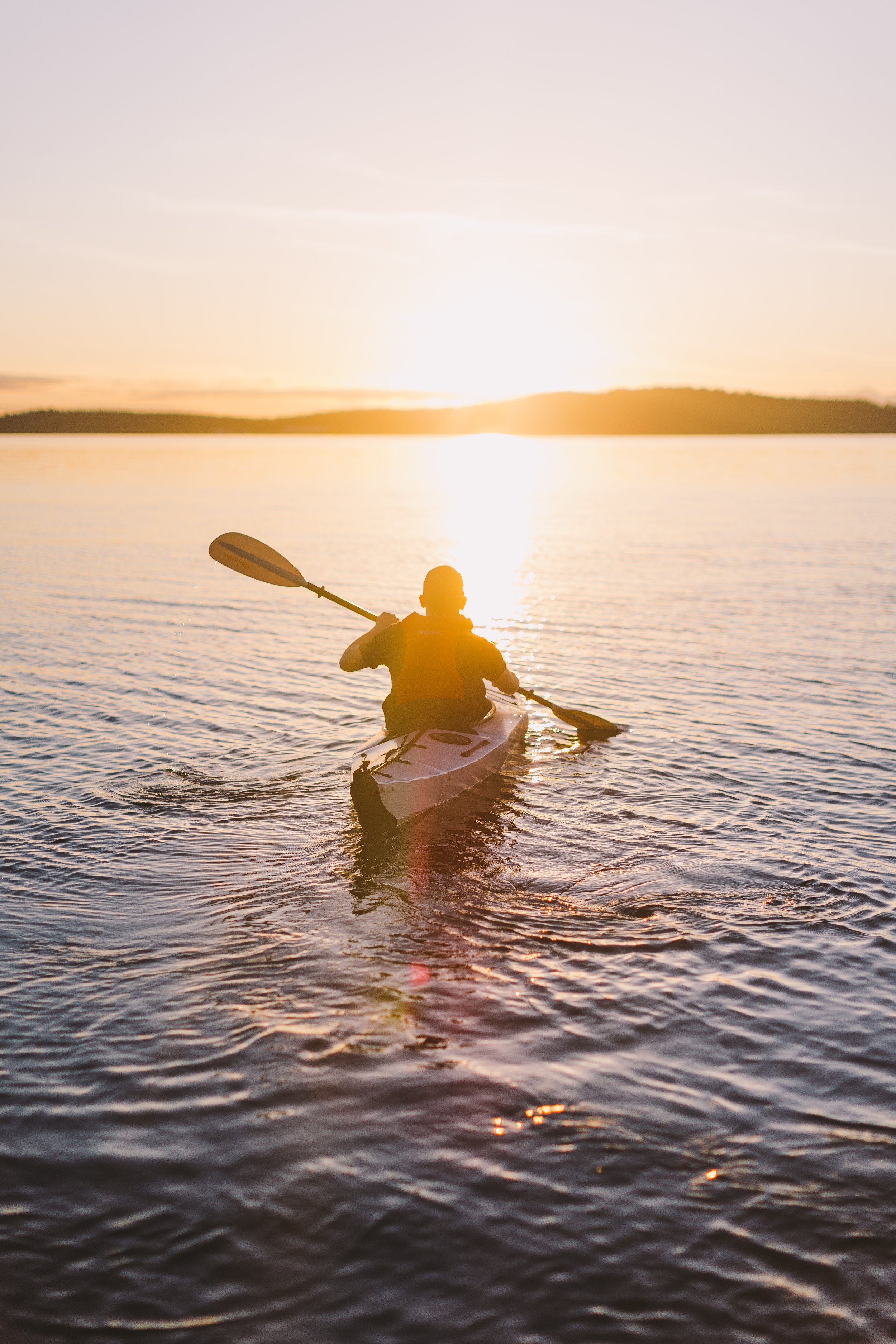 A person kayaking on a calm body of water during sunset, with the sky illuminated in warm tones and a silhouette of land in the background.