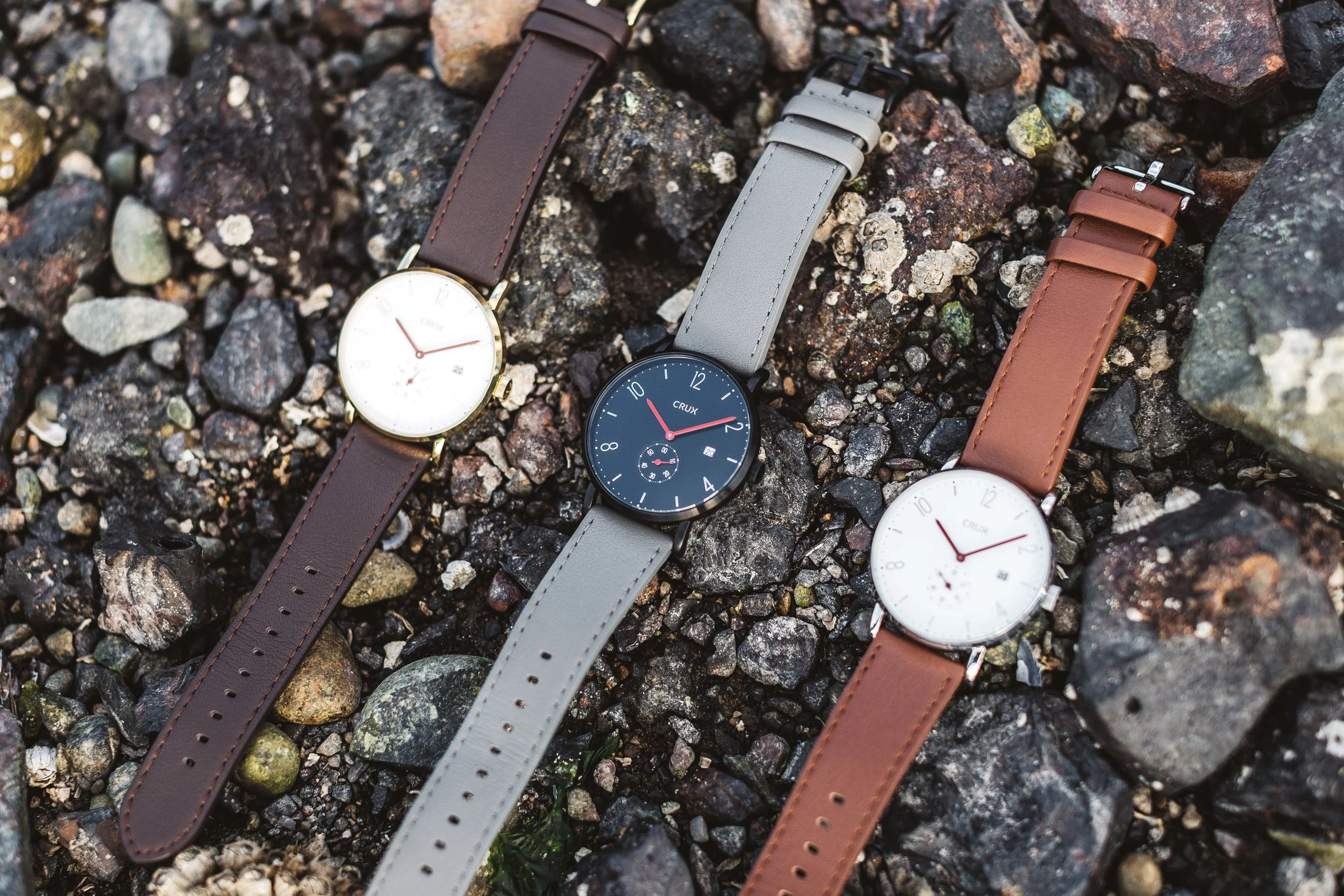 Three wristwatches with leather bands, one with a brown band, one with a gray band, and one with a tan band, lying on a bed of small rocks and gravel.