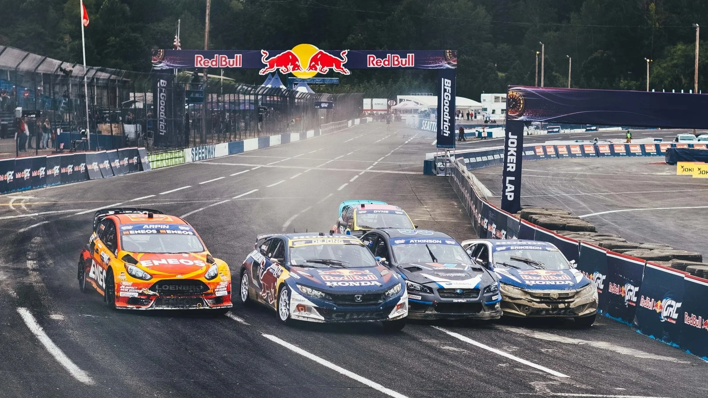 Race cars on a dirt track at a motorsport event, with some cars colliding and kicking up dust, surrounded by barriers with sponsor logos and an overhead banner with the Red Bull logo.
