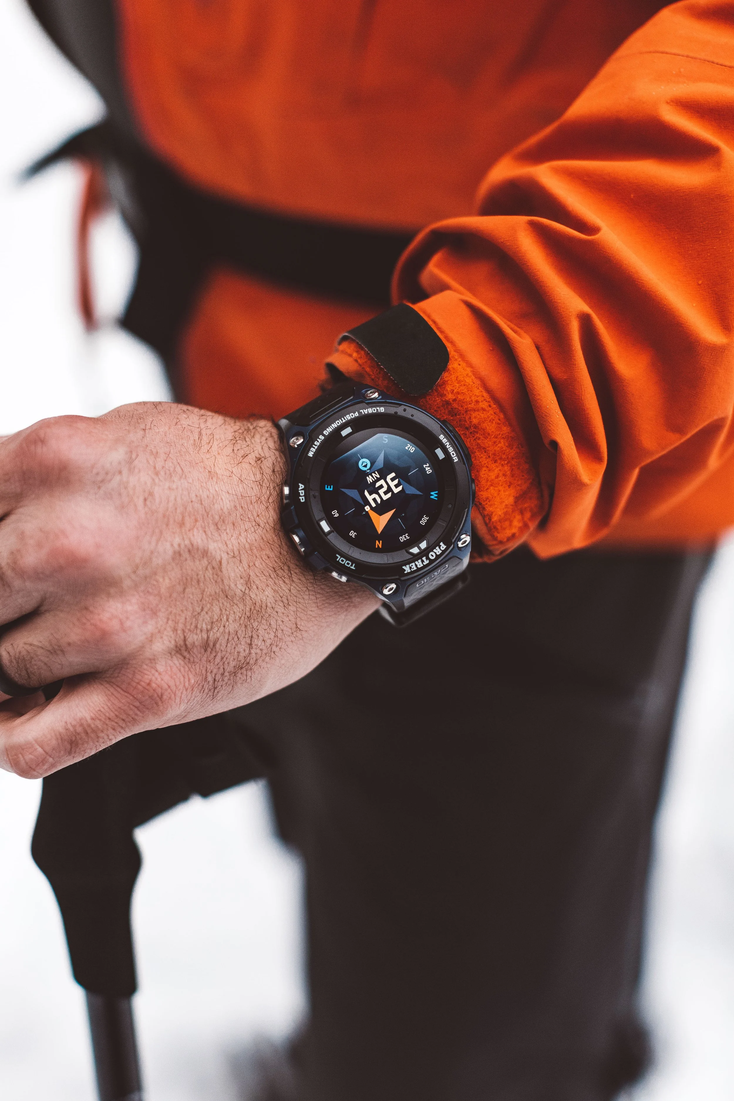 Close-up of a person's left wrist wearing a black outdoor smartwatch with a compass display. The person is dressed in an orange jacket and black pants, and is holding a black walking cane. The background is blurred with white and dark tones.