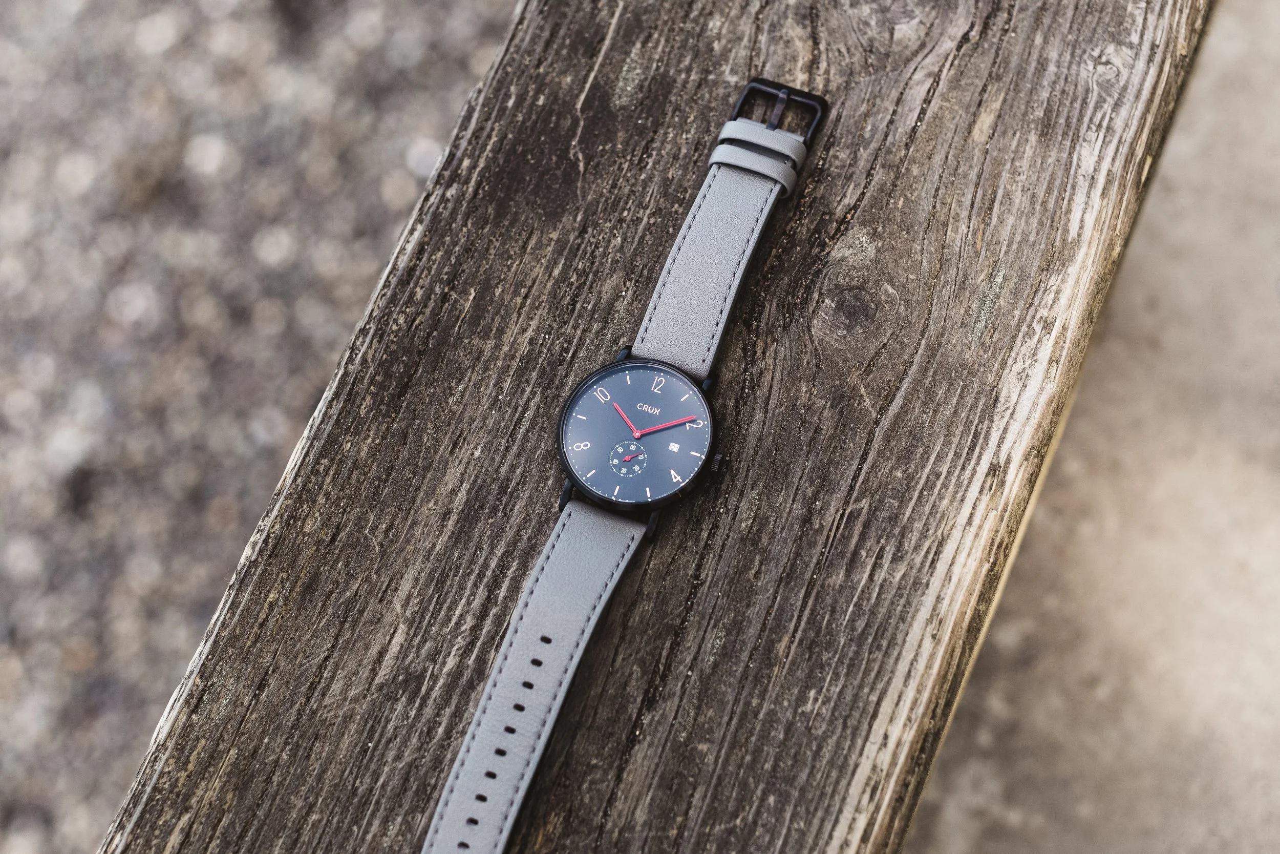 A wristwatch with a black face and red hands, displaying time around 12:09, with a small date window, attached to a light gray leather strap, resting on a weathered wooden surface.